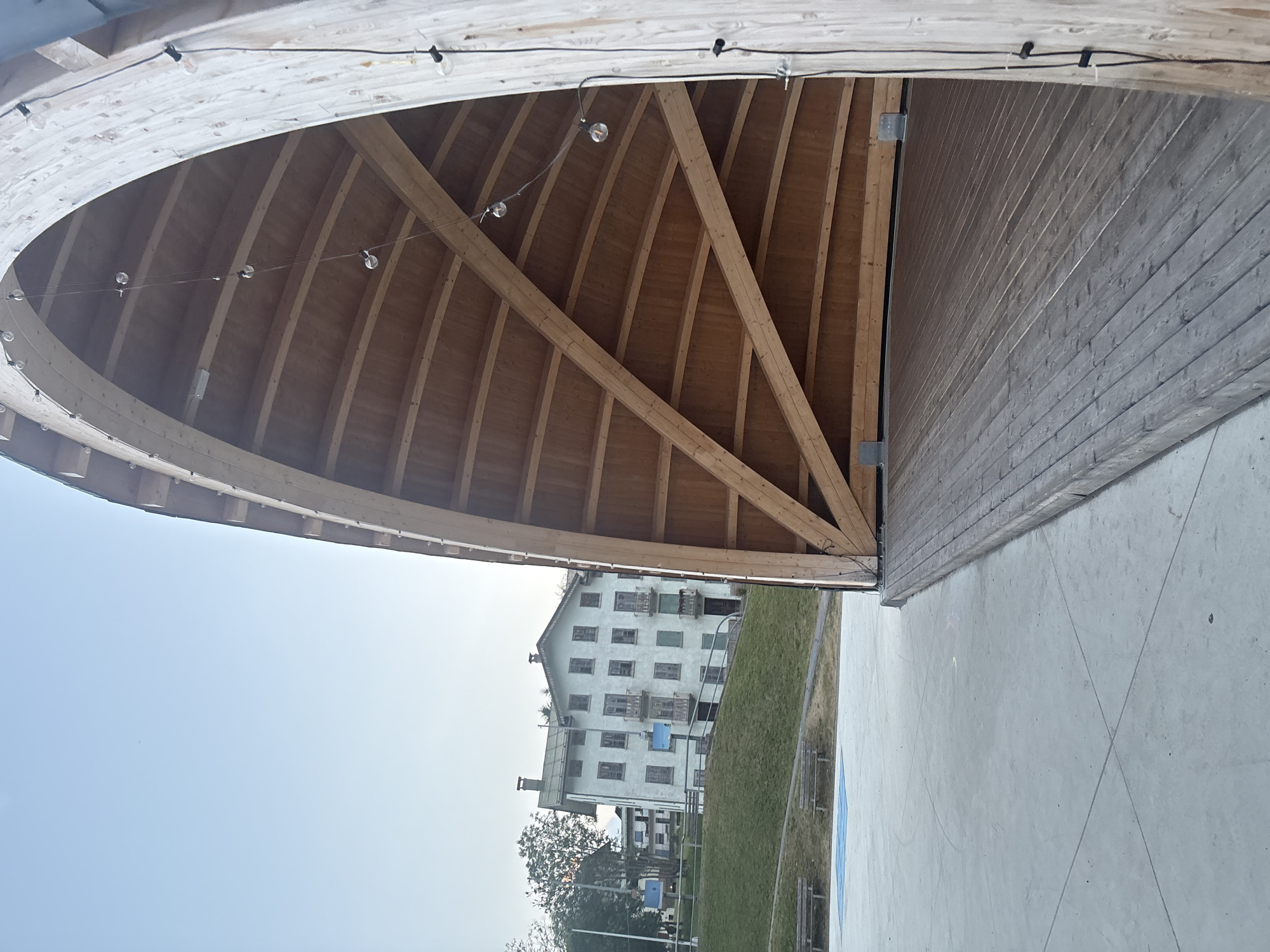 The image depicts an outdoor wooden stage structure with a curved roof supported by large wooden beams. String lights are suspended from the interior of the roof. To the left of the stage, there is a concrete ground area with faint markings. Further in the background, a grassy slope leads up to several buildings, one of which is a multi-story light-colored structure with numerous windows and balconies. Two wooden benches are visible on the grass, and a blue square marking is on the concrete ground. The sky is clear and pale blue.