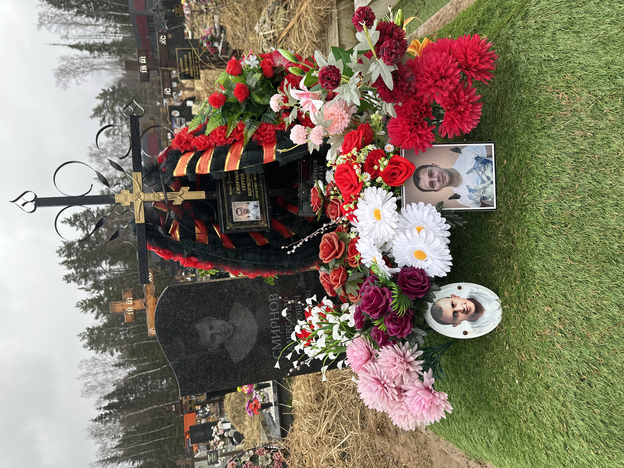 The image depicts a burial site in what appears to be a cemetery, given the presence of multiple tombstones and grave markers visible in the background. The foreground features a dark, polished gravestone adorned with a metal cross and an engraved portrait. Red, pink, and white artificial flowers surround the stone, along with decorative wreaths. Two separate framed photographs are placed among the flowers, one showing a young man wearing a white shirt with a picture of wolves on it, and another in an oval frame depicting an infant. The surrounding area is covered in grass. Based on visual analysis, the location cannot be confirmed as Шарапово, Russia without further information.