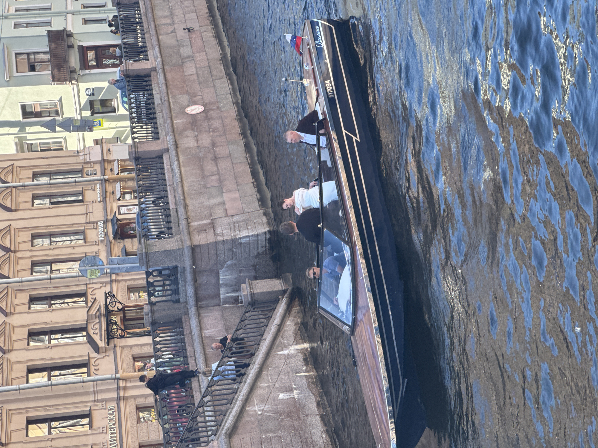 The image shows a contemporary dark blue and wood-trimmed motorboat with a Russian flag on its stern, cruising down a canal in Saint Petersburg, Russia. Four individuals are on board the boat. A man wearing sunglasses and a blue polo shirt is at the helm. Behind him, a woman in a white dress and a man in a black suit are seated. Another man, partially visible from the back, stands at the stern. The boat's hull displays the registration number "517094" and the name "Рислинг" (Risling). In the background, a multi-story building with architectural details and arched windows lines the canal, with people visible on a stone embankment and a staircase leading down to the water. The scene suggests a leisurely ride on a canal, likely a popular tourist route, with historical architecture framing the urban waterway.