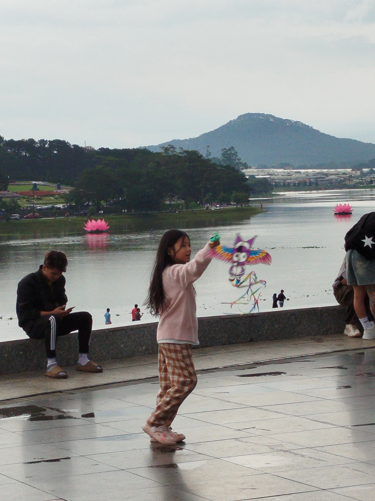 The image captures a scene near Xuan Huong Lake in Da Lat, Vietnam, on what appears to be a cloudy day. In the foreground, a young girl is holding up a colorful kite depicting a cartoon owl. She is wearing a light pink sweater and brown plaid pants. To the left, a young man is seated on a concrete ledge, looking at a mobile phone. He is dressed in dark clothing with Adidas stripes on his pants and is wearing sandals.
In the background, Xuan Huong Lake is visible with floating pink lotus flower decorations. A few people are wading in the shallow water near the edge of the lake. Across the lake, there is a landscaped area with trees and a distant mountain. The ground appears to be wet. Other people can be seen in the background on the right side of the image.