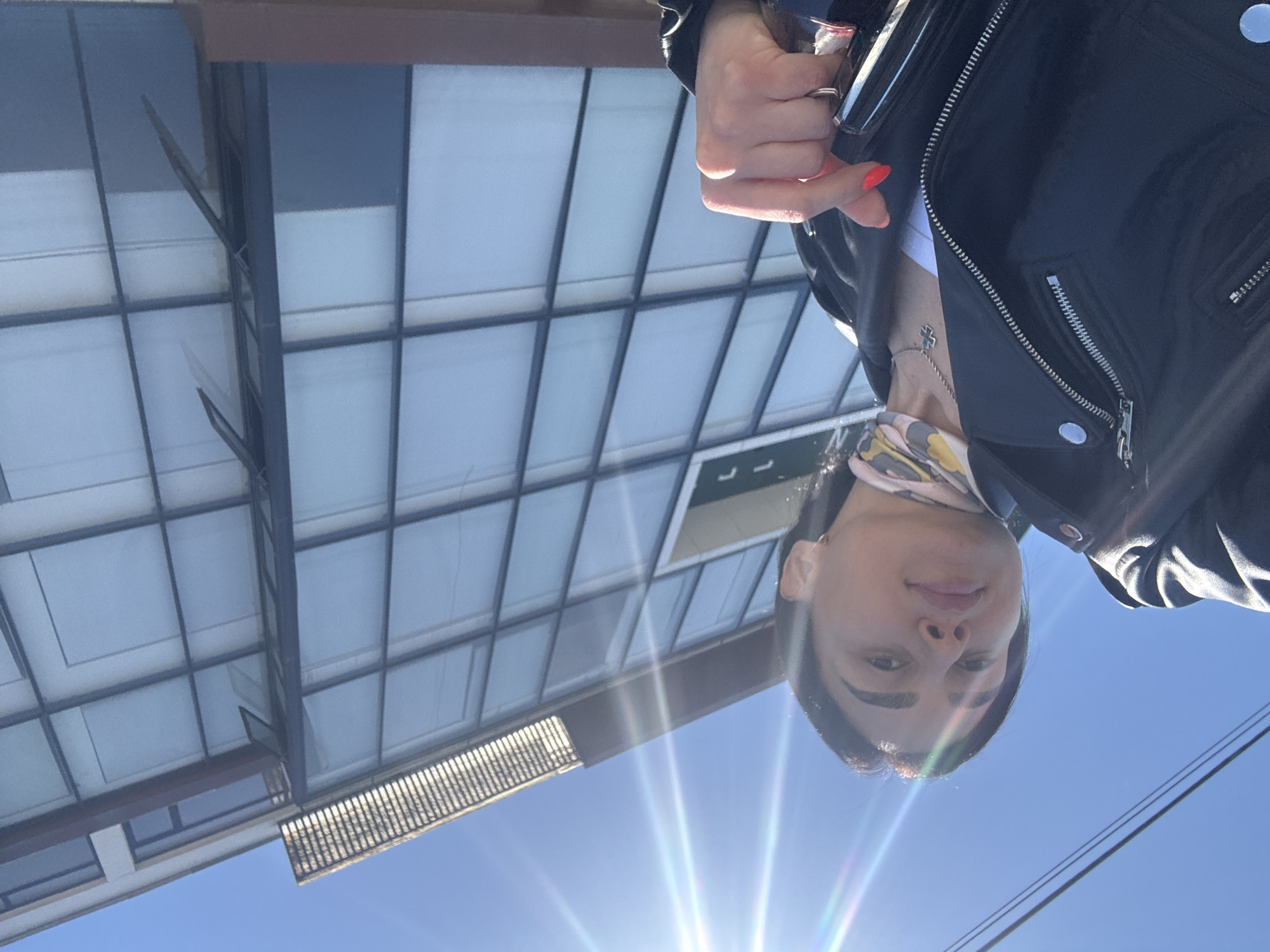 A medium-shot, outdoor, rotated image shows a person wearing a black jacket, a cross necklace, and a patterned scarf. The person's right hand is visible. Behind the person, there is a building with a grid of windows. Above and to the right of the building, the clear, blue sky is visible with some sunlight glares. Power lines are visible in the lower right corner. The location is likely Tbilisi, Georgia.