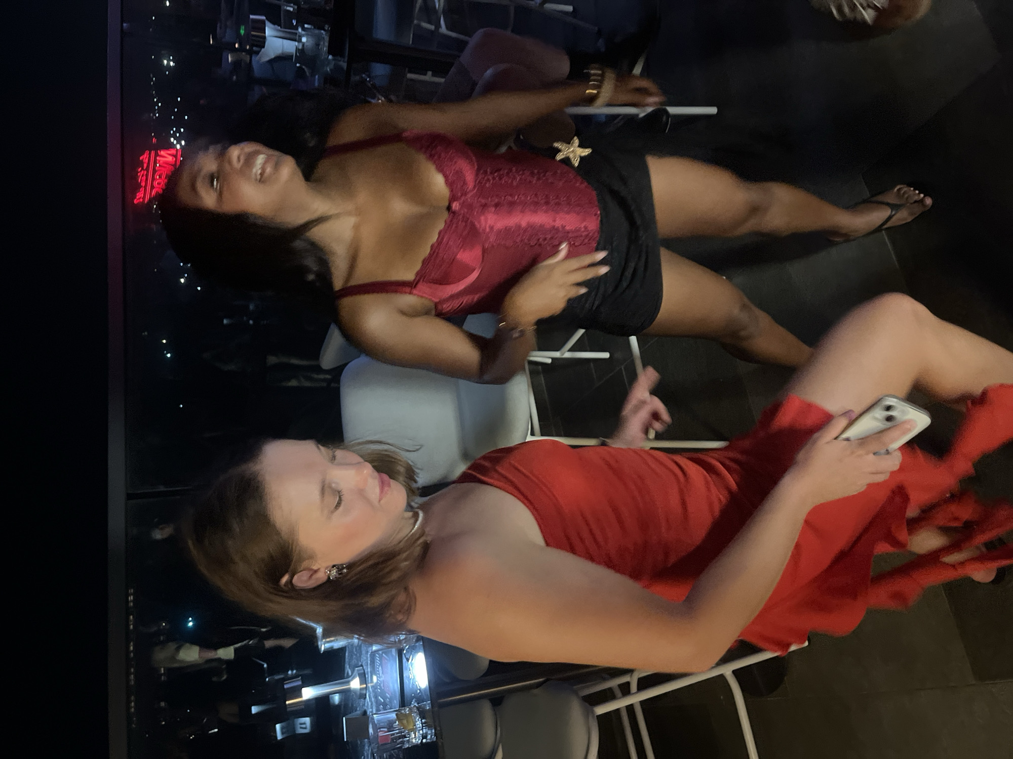 Two women are dancing at night, possibly in a bar or club. The woman on the left wears a red, one-shoulder dress, and the woman on the right wears a red corset top and a black skirt. The background shows a cityscape with lights, suggesting an urban environment. The location is in Da Nang, Vietnam.
