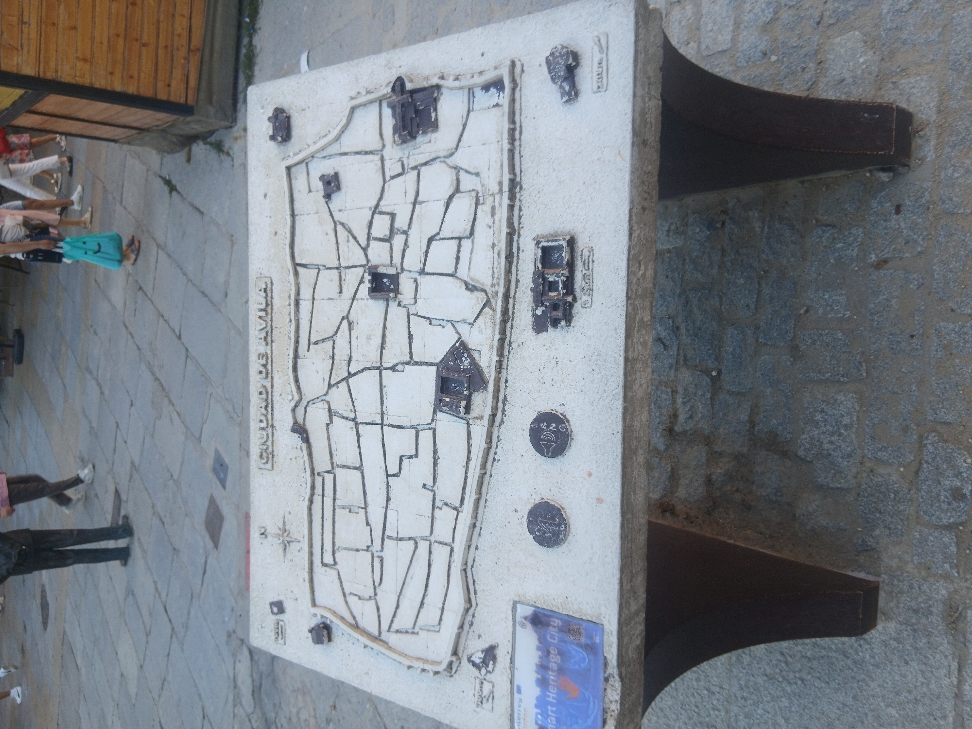 The image shows a relief map of the city of Ávila, Spain, mounted on a stone pedestal in an outdoor public space. The map depicts the city's walls and inner streets, with raised sections representing important buildings or landmarks. Text on the map reads "CIUDAD DE AVILA". To the left of the map, a blue plaque is visible, with partial text including "Smart Heritage City". Two circular medallions, possibly informational markers or decorative elements, are also present on the map's surface. The pedestal is constructed from stone and supported by two dark, curved legs. In the background, paved ground with stone slabs is visible, along with the legs of a few people walking. A wooden structure with a dark roof is located to the right of the scene. The lighting suggests it is daytime.