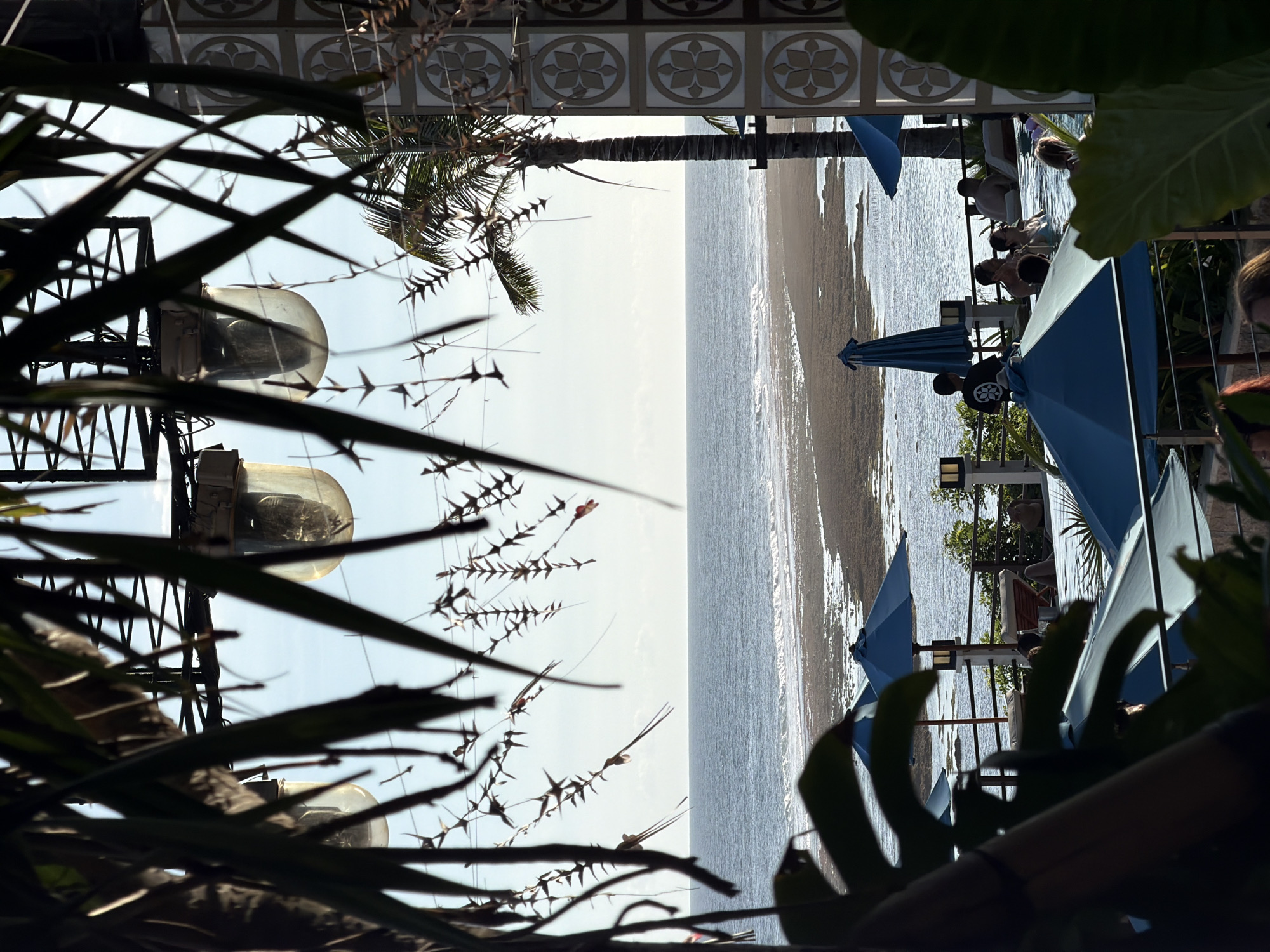 The image depicts a beach scene in Kuta Selatan, Indonesia, possibly viewed from a restaurant or bar overlooking the ocean. In the mid-ground, the ocean meets a sandy beach with several blue umbrellas providing shade to people sitting in beach chairs. The presence of a palm tree suggests a tropical environment. In the foreground, vegetation frames the view on both sides. On the left are some outdoor lighting fixtures, and the bottom and right of the frame have large leafy tropical plants. The composition suggests a vantage point from an elevated position, looking down towards the beach. People are visible under the umbrellas. The overall environment appears relaxed and beach-oriented.