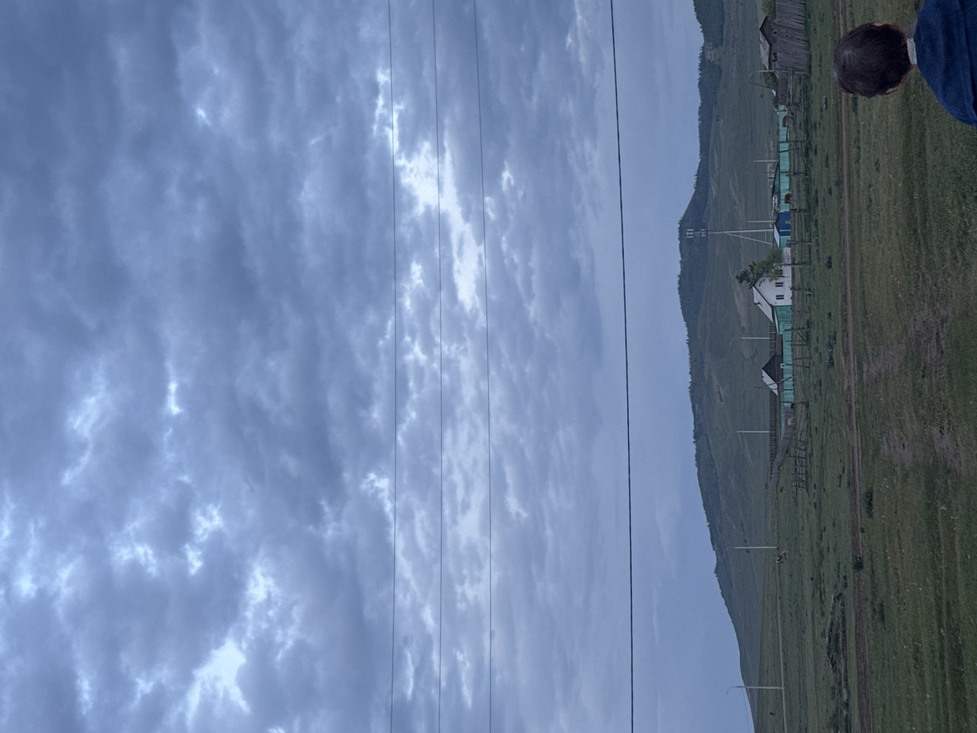 The image shows a view of Нижний Бургалтай, Russia, under an overcast sky with layered gray and white clouds. Power lines are visible, running vertically across the frame. The landscape includes grassy fields, hills, and scattered buildings with green and white paint, some of which have wooden fences. A person's head is visible in the upper right corner.