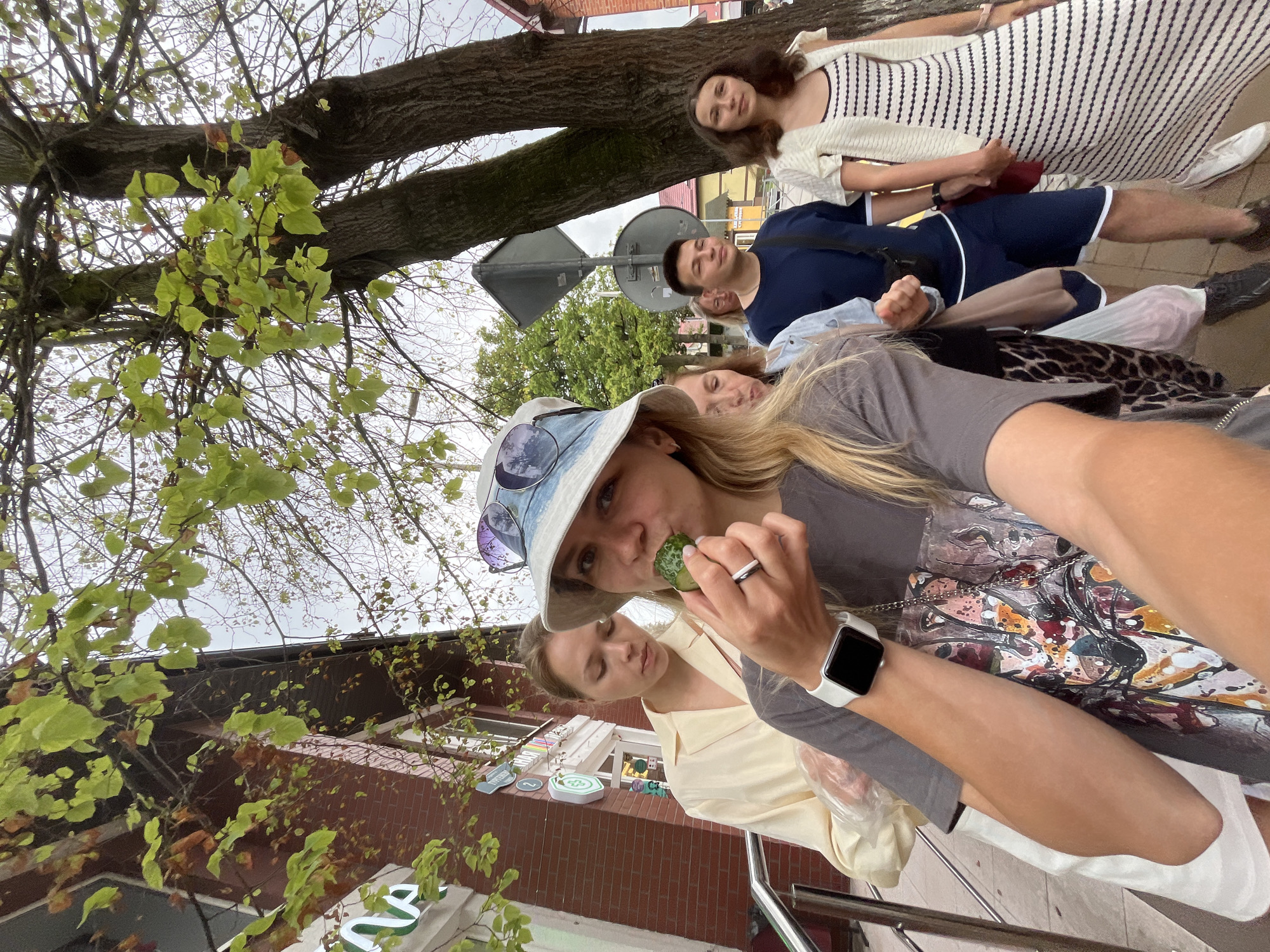 In Zelenogradsk, Russia, a group of people stand on a sidewalk near a brick building and a tree. A woman in the foreground, wearing a bucket hat and sunglasses, is taking a selfie while eating a piece of cucumber. Behind her, a woman in a yellow shirt and another in a light blue shirt are visible. To the right, a man in a blue shirt and shorts stands next to a woman in a striped dress and white cardigan. The environment suggests an outdoor urban setting with clear skies.