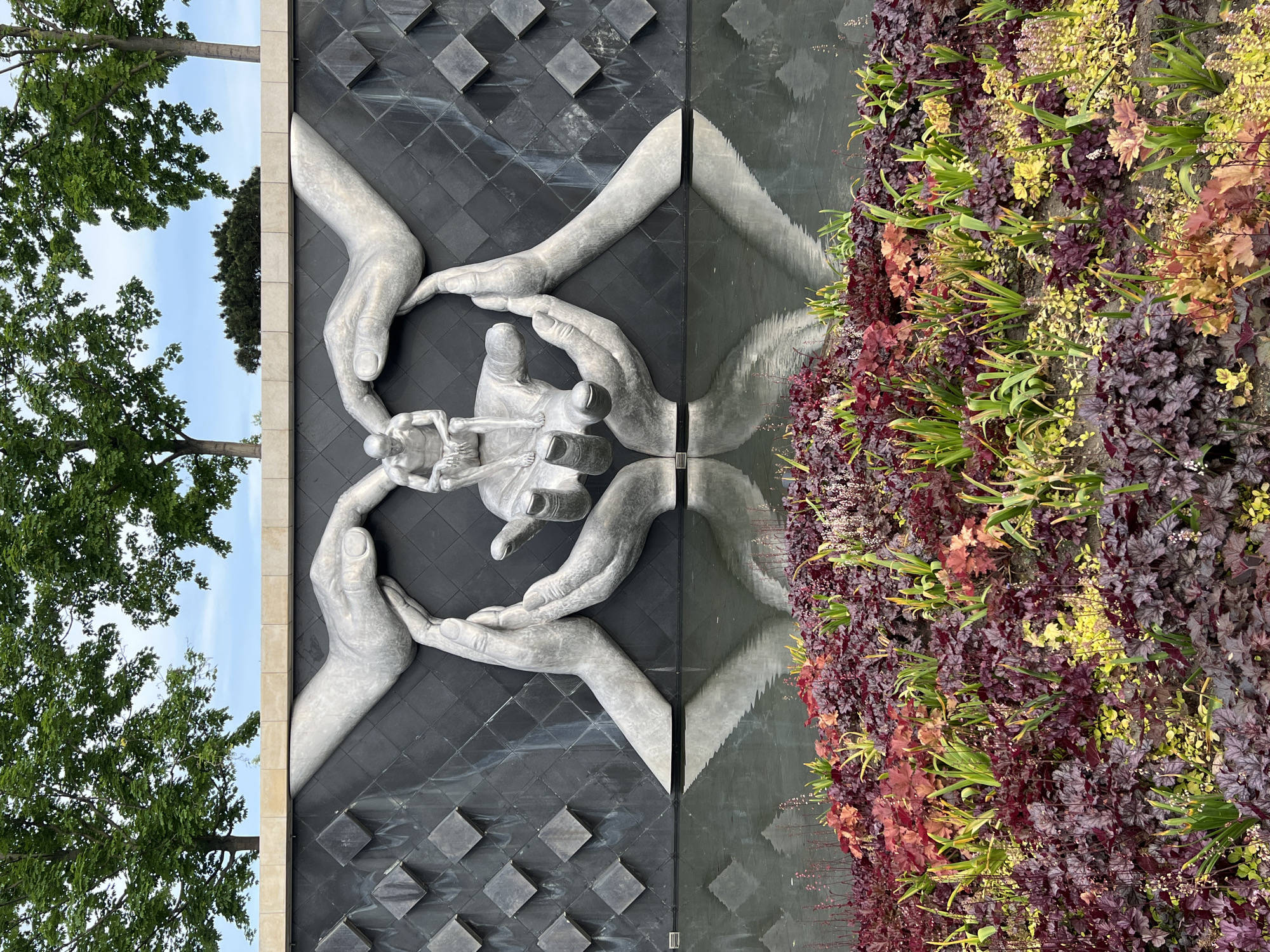 The image shows a fragment of a building facade in Krasnodar, Russia, featuring a dark grey, diamond-patterned wall with a silver bas-relief depicting four large hands cradling a group of human figures. To the right, a wall covered in dense foliage of various colors, predominantly reds, purples, and yellows, vertically borders the textured wall. The wall also reflects an image of the stone hands to the right side. A tree is visible in the sky on the far left, and the image is taken in daylight.