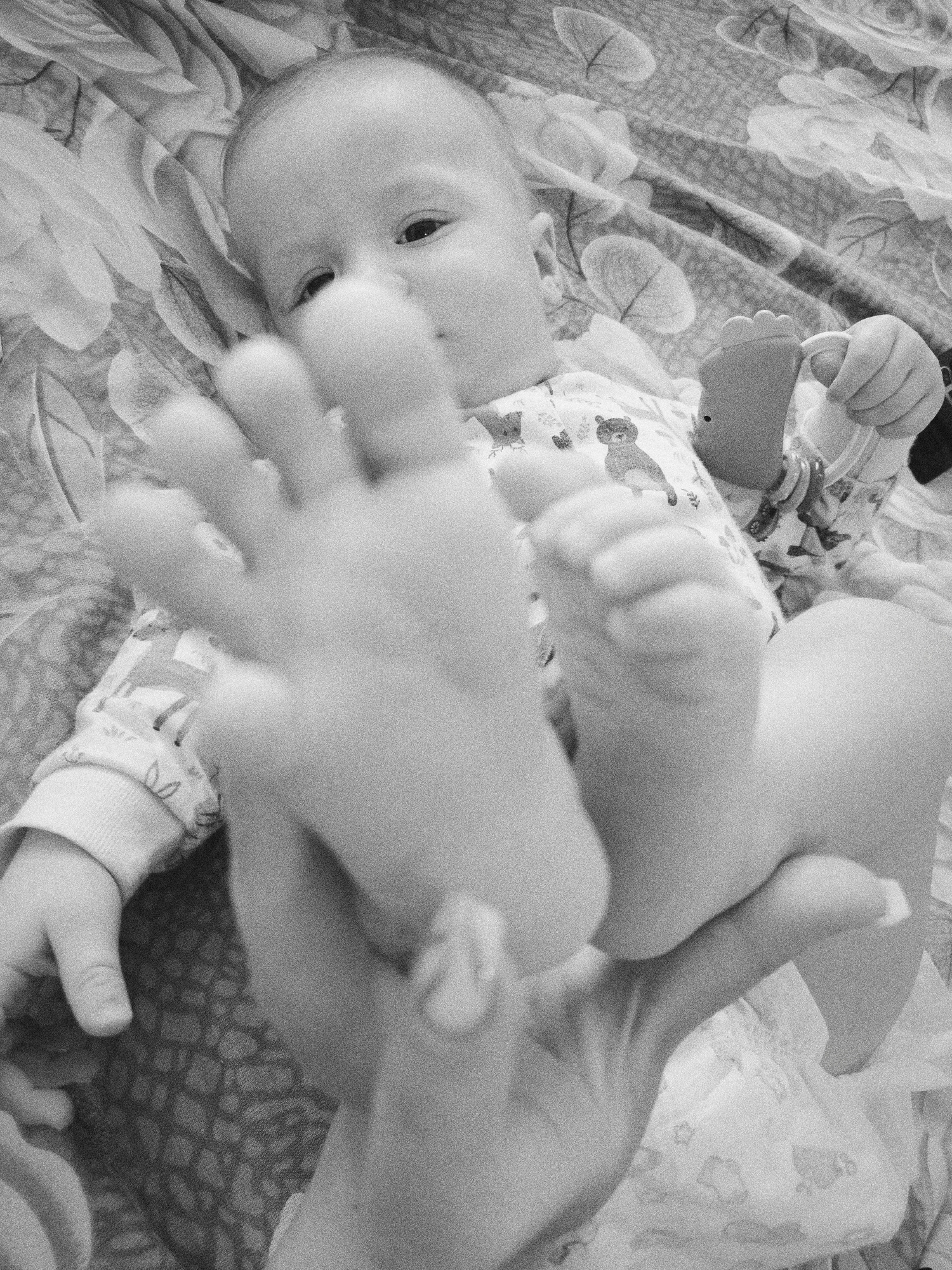 A black and white, high-angle photograph shows a baby lying on a patterned blanket. The baby, with a bald head, looks toward the camera with a relaxed expression. The baby is wearing a long-sleeved garment with an animal print design. An adult's hand holds the baby's hand, obscuring part of the baby's face. The baby's other hand holds a toy in the shape of a foot. The baby's legs are raised, and part of what appears to be a diaper is visible. There is no location data provided.