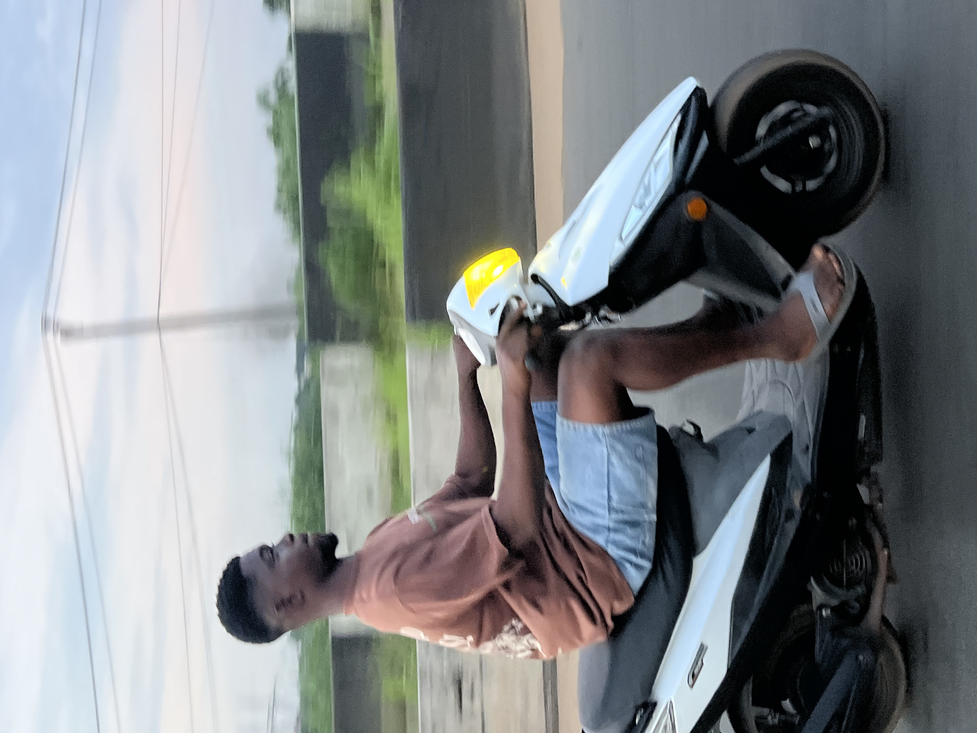 A young Black man is riding a white scooter in Orifite, Nigeria. He has a short, faded haircut and is wearing a brown t-shirt and light blue denim shorts. The scooter has a yellow headlight. The background shows a blurred landscape with greenery and power lines against a cloudy sky, suggesting an outdoor, possibly semi-urban or rural environment. The scooter is in motion on a paved surface, indicated by the motion blur.