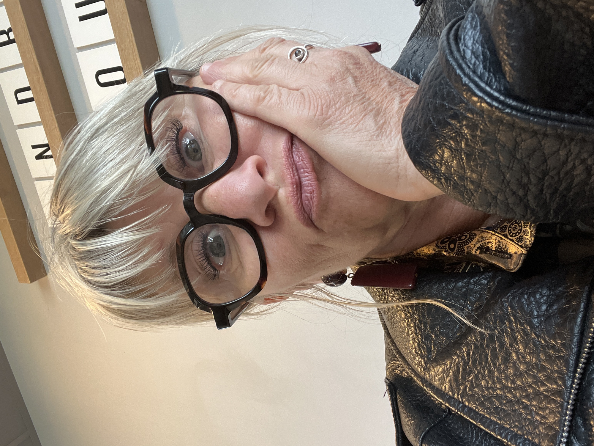 A person with white hair is facing the camera, wearing eyeglasses, a ring, and a black leather jacket. The person's right hand is touching their forehead. In the background, there is a set of white panels with black lettering. The location is unknown, and there is no indication that this image was taken in Andrézieux-Bouthéon, France.