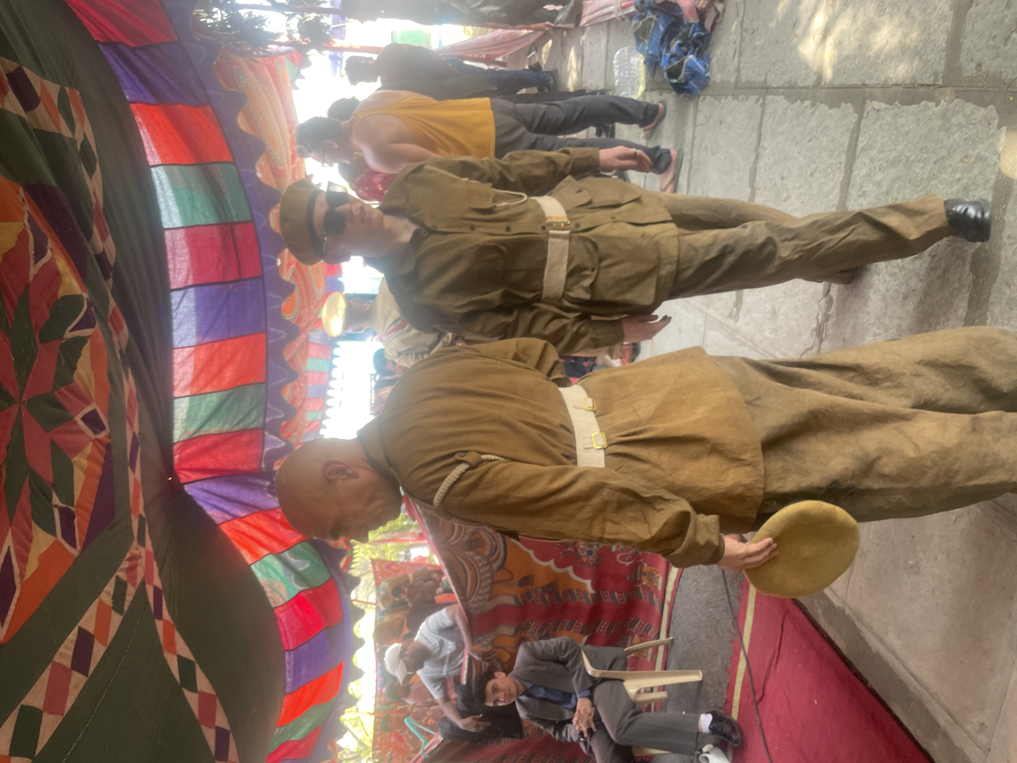 The image captures a scene in Anajpur, India. Underneath a vibrant, patterned tent canopy, two individuals are dressed in tan uniforms with white belts and hats. The foreground figure, bald, holds a tan object in his hand. Behind them, a person in a yellow tank top is visible, along with another in a suit sitting on a chair. The ground is a mix of stone pavement and a red rug. Other people can be seen in the background, some seated and some standing.