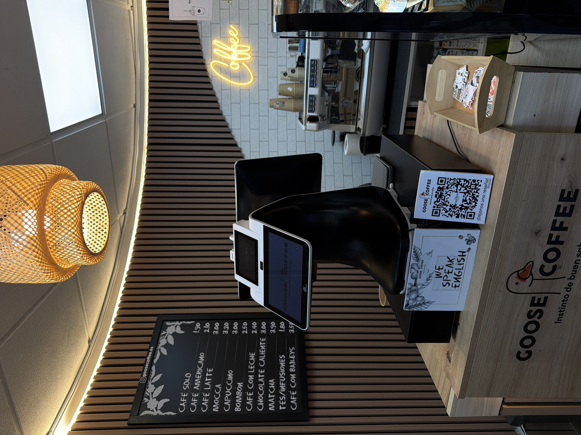 Here is a detailed description of the image:
**Location:** The image appears to be taken inside a Goose Coffee establishment, possibly in Valencia, Spain, given the reference to "@goosecoffee.es" and "Instinto de buen sabor" which translates to "Instinct of good taste" in Spanish.
**Scene:** The scene is a view of the front counter area of the cafe. Key elements include:
* **Menu Board:** A chalkboard-style menu lists coffee and drink options with their prices in euros, such as "CAFÉ SOLO" (1.90), "CAFE AMERICANO" (2.10), and "CAFE CON BAILEYS" (3.50). The Instagram handle "@goosecoffee.es" is also displayed.
* **Point of Sale System:** A digital point of sale system with two screens is visible. One screen displays the Goose Coffee logo, suggesting it is a customer-facing screen.
* **Signage:** Various signage elements are present:
* A sign with "WE SPEAK ENGLISH" displayed, indicating the staff's language capabilities.
* A QR code sign to provide a review.
* The "GOOSE COFFEE" logo.
* **Coffee Machine:** A professional coffee machine is visible in the background, suggesting the coffee preparation area.
* **Wooden Decor:** Wood is used for the wall paneling, counter, and box, providing a warm aesthetic.
* **Light Fixture:** A woven, rattan-style light fixture is visible on the left.
* **Neon Sign:** A neon sign that says "Coffee" is above the coffee machine.
* **Stickers:** Stickers are displayed on a wooden tray next to the point of sale.
**Subjects:**
* The primary focus is on the coffee shop's setup and brand presentation, rather than on any human subjects.
**Objects:**
* Coffee menu board, POS system, coffee machine, wooden counter, lighting fixture, neon sign, sticker tray.
**Actions/Interactions:**
* The image suggests the cafe is open and ready to serve customers.
**Environment:** The cafe environment appears modern and well-maintained, aimed at providing a welcoming experience.