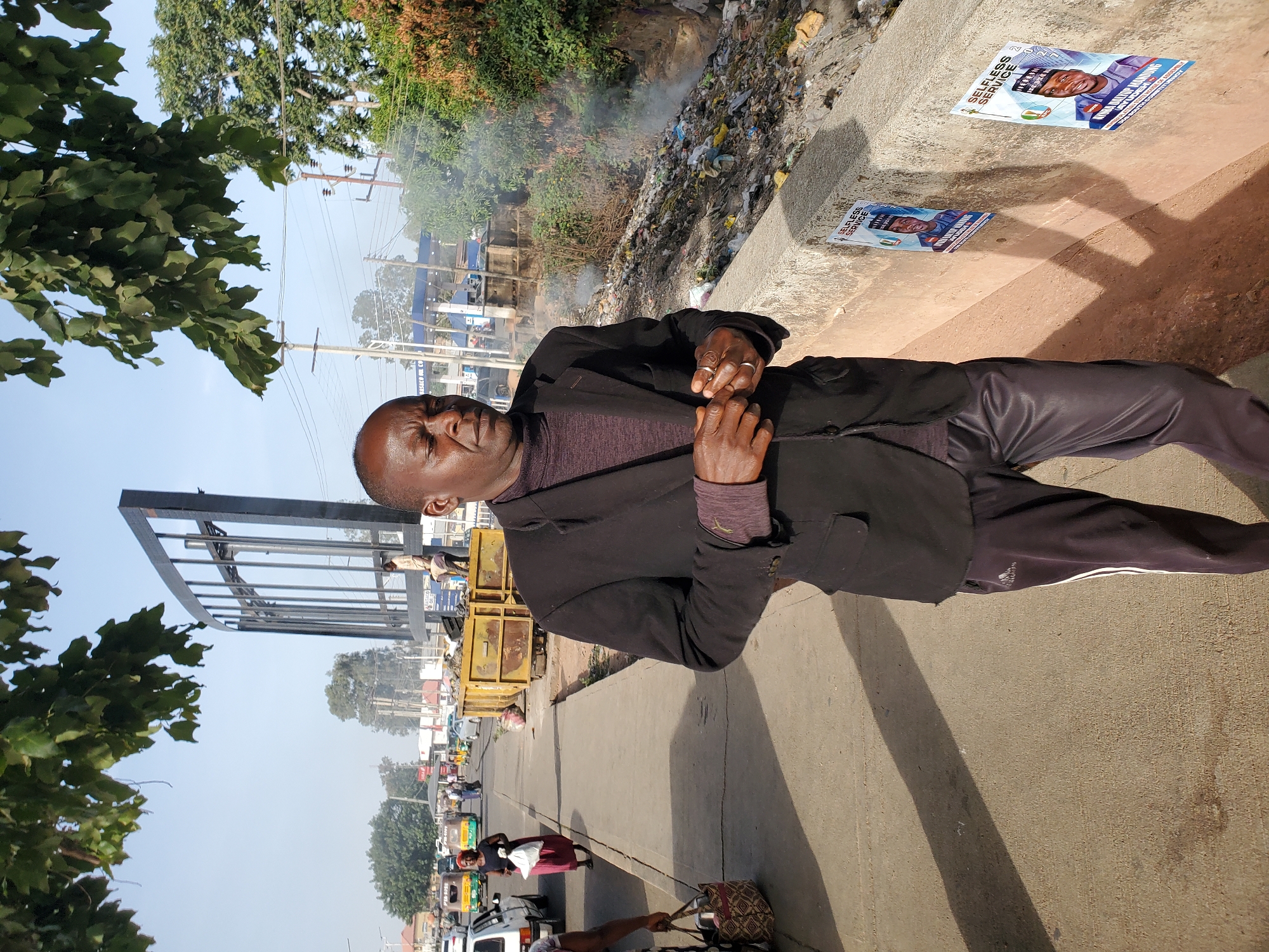 This image captures a vibrant street scene, likely in Jos, Nigeria, showcasing a blend of urban activity and environmental challenges. The central figure is a middle-aged man, positioned in the foreground on a concrete sidewalk. He is dressed in a dark blazer over a purple long-sleeved shirt and dark trousers with a white stripe down the side, bearing a faint Adidas logo. His hands are clasped in front of him, displaying rings on one finger, and he maintains a neutral or slightly serious expression.
Behind the man, across a concrete barrier, a substantial pile of refuse is visibly burning, emitting plumes of smoke into the air. This large accumulation of various types of waste forms a stark backdrop. Attached to the concrete barrier are two blue political posters, both featuring a smiling man in traditional attire. Prominently displayed text on these posters reads "SELFLESS SERVICE" and "MUSA DANIEL LANDING," with "2023" also visible, indicating an election context.
To the man's left, the street is bustling with activity, featuring several vehicles, including cars and auto-rickshaws (Keke Napep), along with other pedestrians. In the mid-ground, a large yellow skip or dumpster is visible. The scene is framed by lush green trees on both sides, while overhead, power lines and utility poles crisscross the sky, characteristic of an urban setting. An empty, large metal billboard structure stands on the far left. The bright sunlight casts clear shadows on the ground, suggesting it is mid-morning or afternoon under clear weather conditions. The overall impression is one of everyday life in a developing urban area, highlighting community activities alongside visible environmental issues such as waste management.