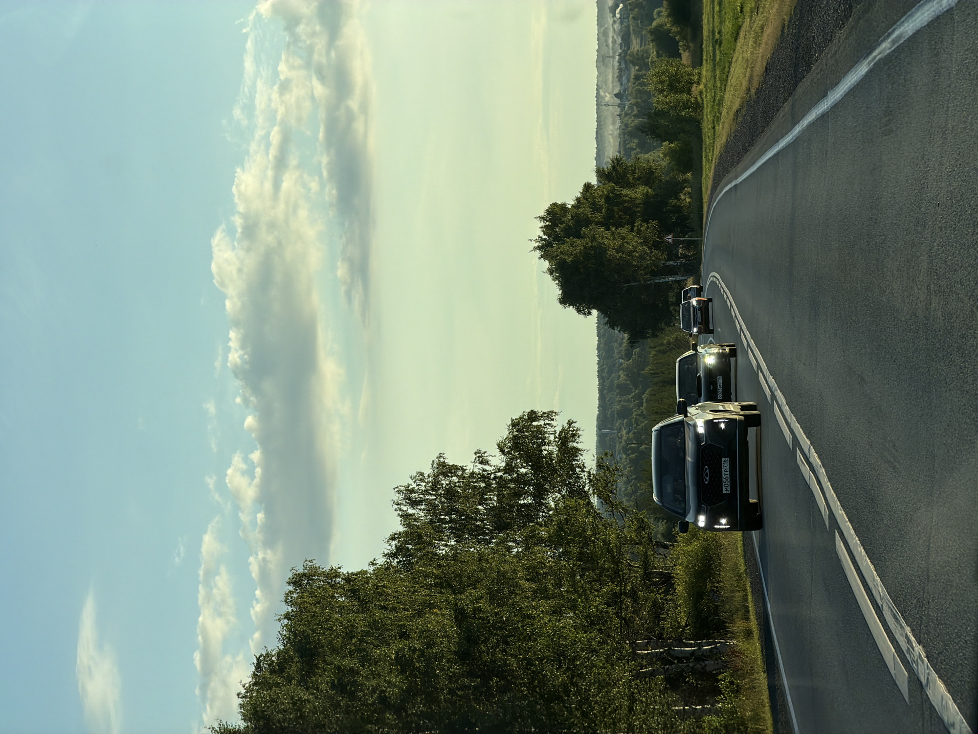 A group of cars are driving on a rural road in Kulaevo, Russia. The lead vehicle, a dark-colored SUV, is in the foreground, its headlights illuminated. Behind it are several other cars, also on the road. The road is paved and has white lane markings. Lush green trees line the left side of the road, and a rolling landscape with fields and scattered trees stretches into the background. The sky above is a pale blue with scattered white clouds, suggesting it is either early morning or late afternoon. The scene appears peaceful and serene.