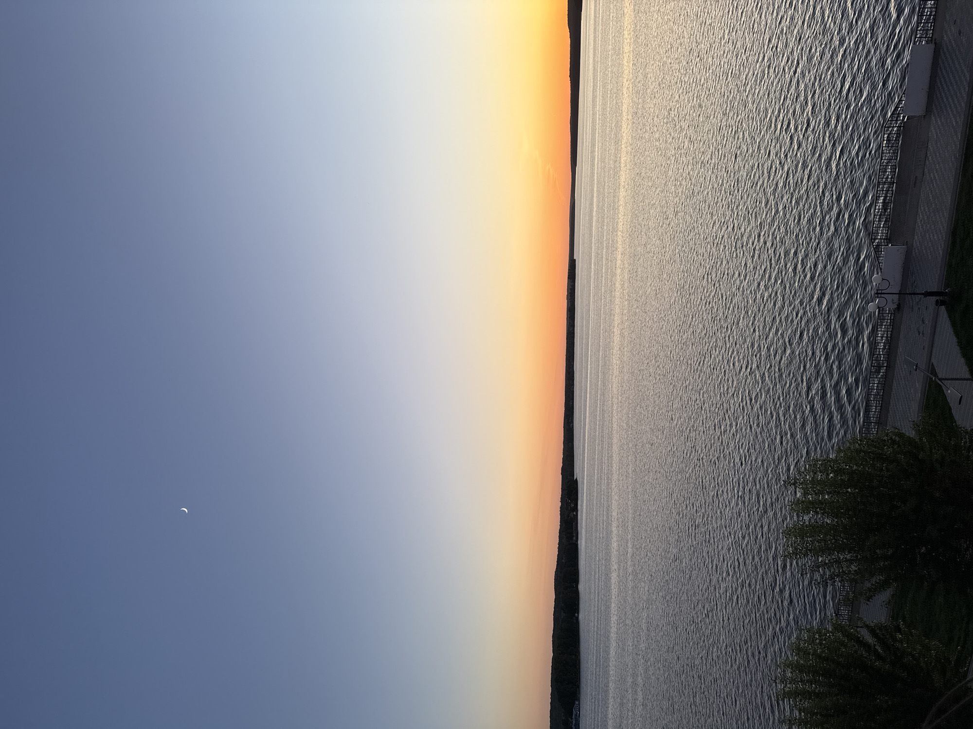 The image depicts a waterfront scene in Votkinsk, Russia, likely taken near sunset. The sky transitions from blue to orange, reflected in the water. A crescent moon is visible in the blue portion of the sky. The water is textured with ripples, reflecting the fading sunlight. A waterfront promenade with railings, concrete, and grass is visible on the right side of the image. Trees partially obscure the bottom right corner.