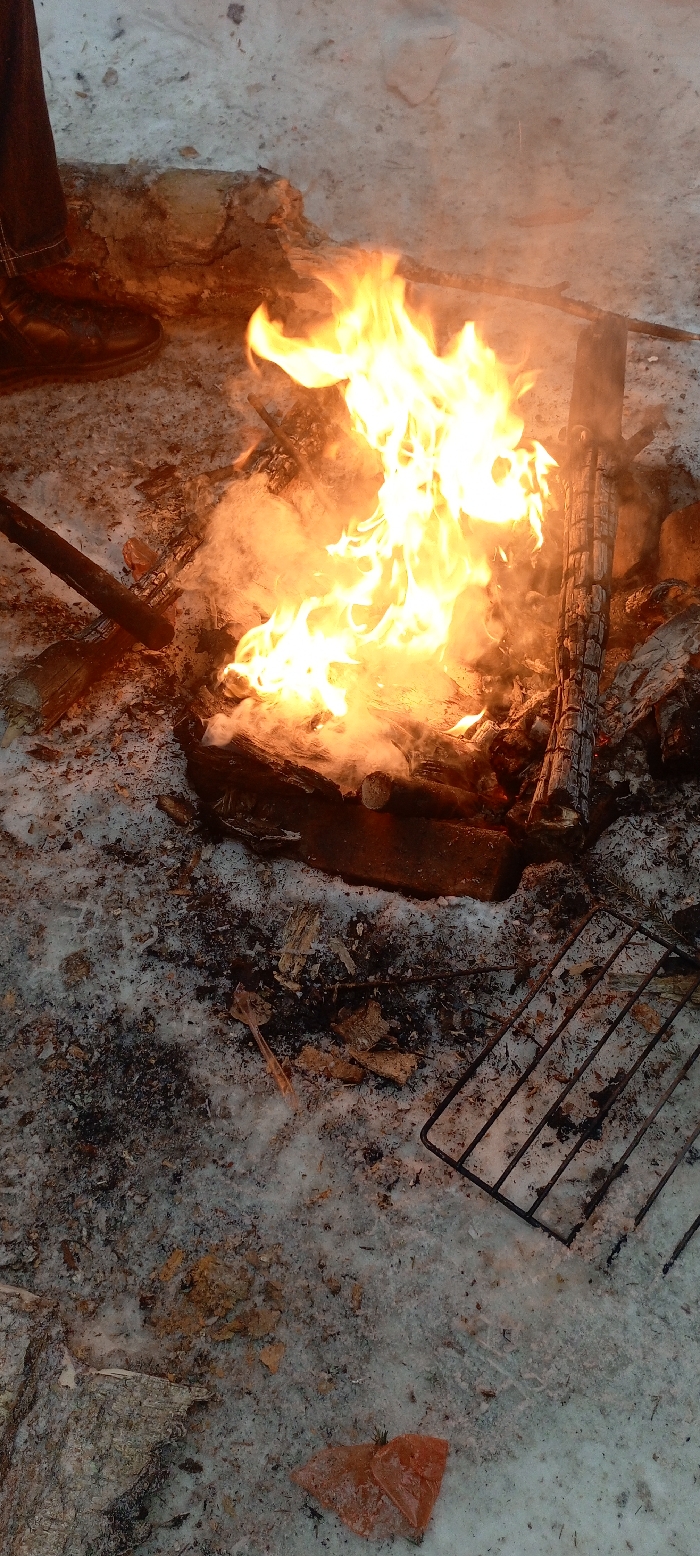 An outdoor scene captured from an elevated perspective depicts a wood-burning campfire on snow-covered ground within the city of Unknown, Unknown. Bright yellow and orange flames with white smoke rise from a pile of burning logs and wood debris in the central-right portion of the frame. The immediate ground around the fire consists of dirty, melting snow, dark ash, and scattered wood fragments. In the upper left quadrant, the lower portion of a person's leg, clad in dark pants and a dark boot, is visible near the fire. A rectangular black metal grill grate lies on the snow in the lower right. A translucent plastic utensil (possibly a spoon or scoop) and a crinkled piece of orange material are present on the snow in the lower foreground.