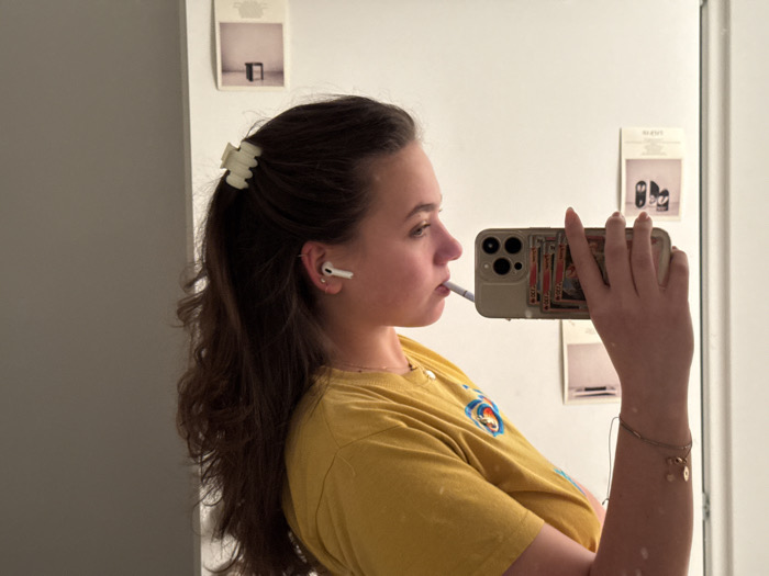 A woman takes a mirror selfie. She's wearing a yellow shirt, has her brown hair partly up with a cream claw clip, and has AirPods in her ears. She holds a phone with playing cards in its clear case. In her mouth is a cigarette. The mirror reflects several framed images on the wall behind her. She wears a gold charm bracelet.