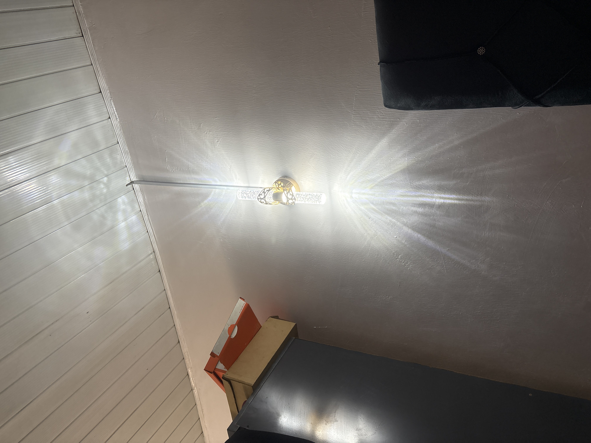 The image depicts an indoor scene with a ceiling that appears to be a combination of painted drywall and wood paneling. A light fixture, illuminated and causing significant glare, is mounted on the drywall portion of the ceiling. Near the bottom of the image, there are cardboard boxes and a dark piece of furniture. The presence of the furniture suggests the scene is within a living space. The image does not contain information about people or actions, and there is no verifiable information about the location within Gura Top, Nigeria.