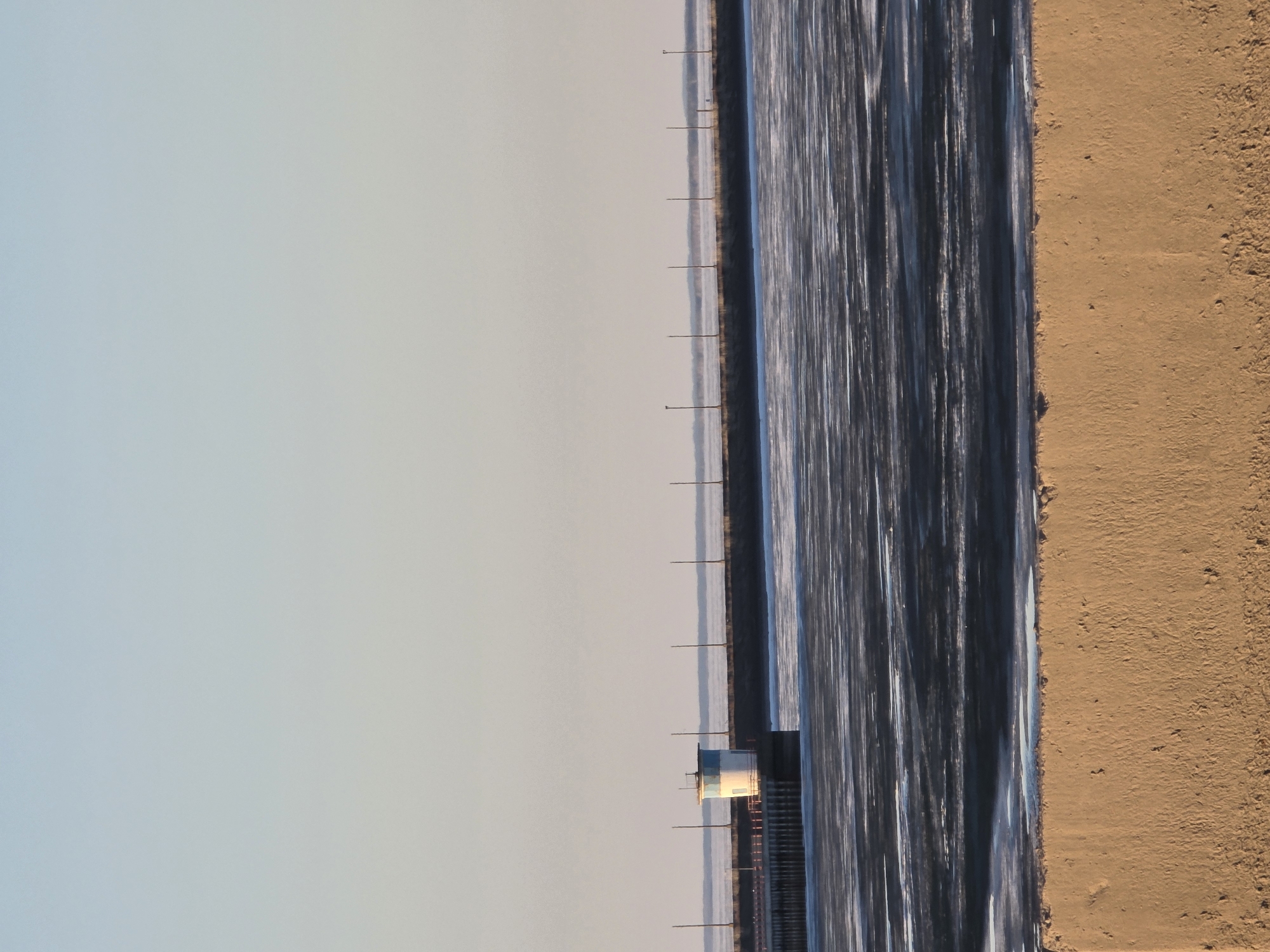 The image depicts an aerial view of what appears to be a body of water, possibly a bay or lake, situated next to a sandy shore. A narrow structure with a small building is located along the water's edge, supported by posts. The water surface shows distinct patterns, possibly indicative of currents or surface texture. The sandy area adjacent to the water is uniform in tone. The image lacks any human presence or movement. The location is Chaikovsky, Russia.