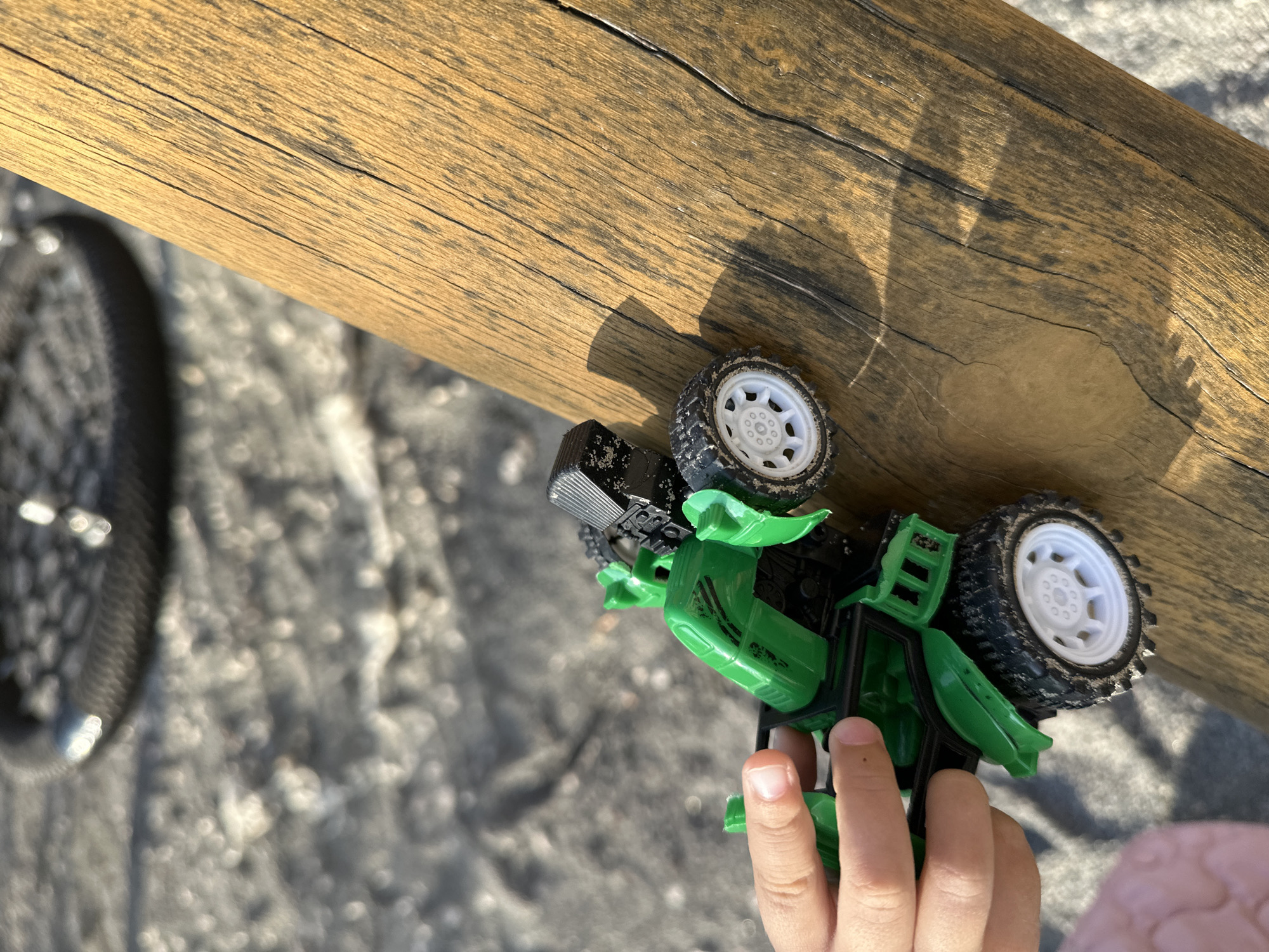 A person's hand holds a green and black toy car on a wooden surface. The car has four white-rimmed wheels with black tires. The background is out of focus and appears to be sandy ground with a large, dark, circular object partially visible, possibly a tire. The wooden surface has prominent grain and cracks, with a shadow of the toy car cast upon it. The scene is outdoors, suggesting it may be a park or playground.