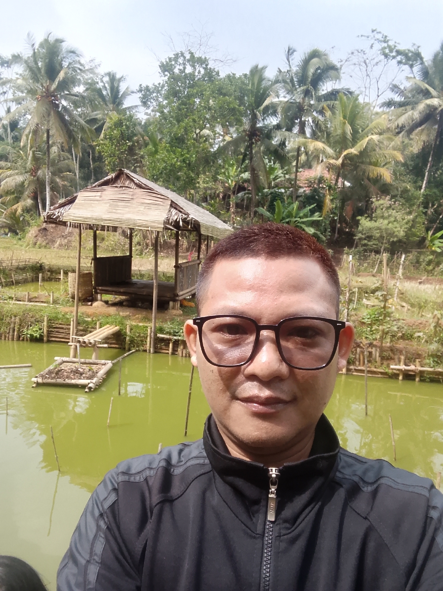 A man wearing glasses and a black jacket poses for a selfie in front of a pond in Cikukulu, Indonesia. The pond is greenish-yellow, and wooden stakes protrude from the water. To the left of the pond, a small raft made of bamboo is floating. Behind the pond is a rustic hut with a thatched roof, built on stilts. The background is filled with lush greenery, including numerous coconut trees, and a bright sky. The man's expression is neutral.