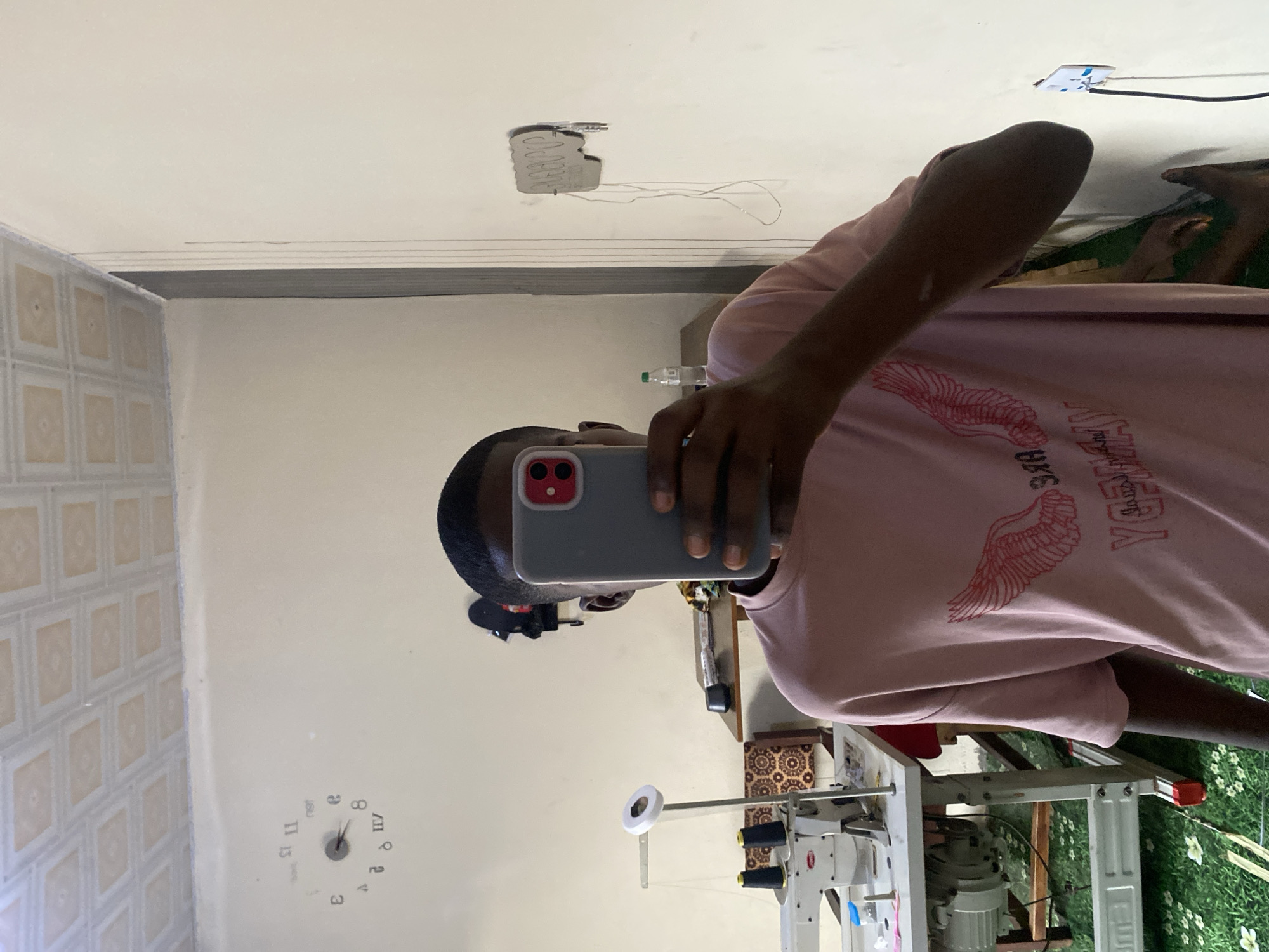 A young Black male is taking a selfie in a room. He is wearing a pink t-shirt with a wing design and is holding a gray smartphone. In the background, there is a sewing machine, spools of thread, a clock on the wall, and a wall outlet with a plug. The room appears to be a workshop or a living space. There is no indication that this location is in Jauro Yinu, Nigeria.