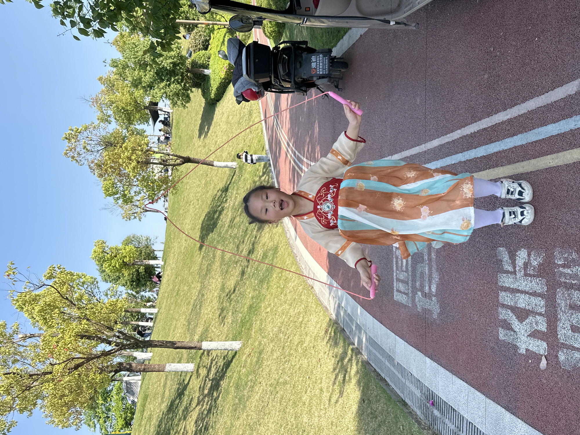 A young girl in a traditional Chinese dress jumps rope on a red path marked with white lines. Her attire consists of an off-white top with red and gold accents, and a flowing skirt with orange, brown, and teal segments. She wears light purple leggings and athletic shoes. She has the jump rope above her head and an expression of focus or determination.
The path runs alongside a grassy area with trees. A white-painted stripe marks the bottom of the trees. To the side of the path is a gray concrete curb and a drainage grate. In the background, there is a moped or scooter parked partially on the path. There are other people in the distance.
The image was taken in Jiangjin, China, likely in a park or recreational area, as implied by the manicured grass, path, and trees.