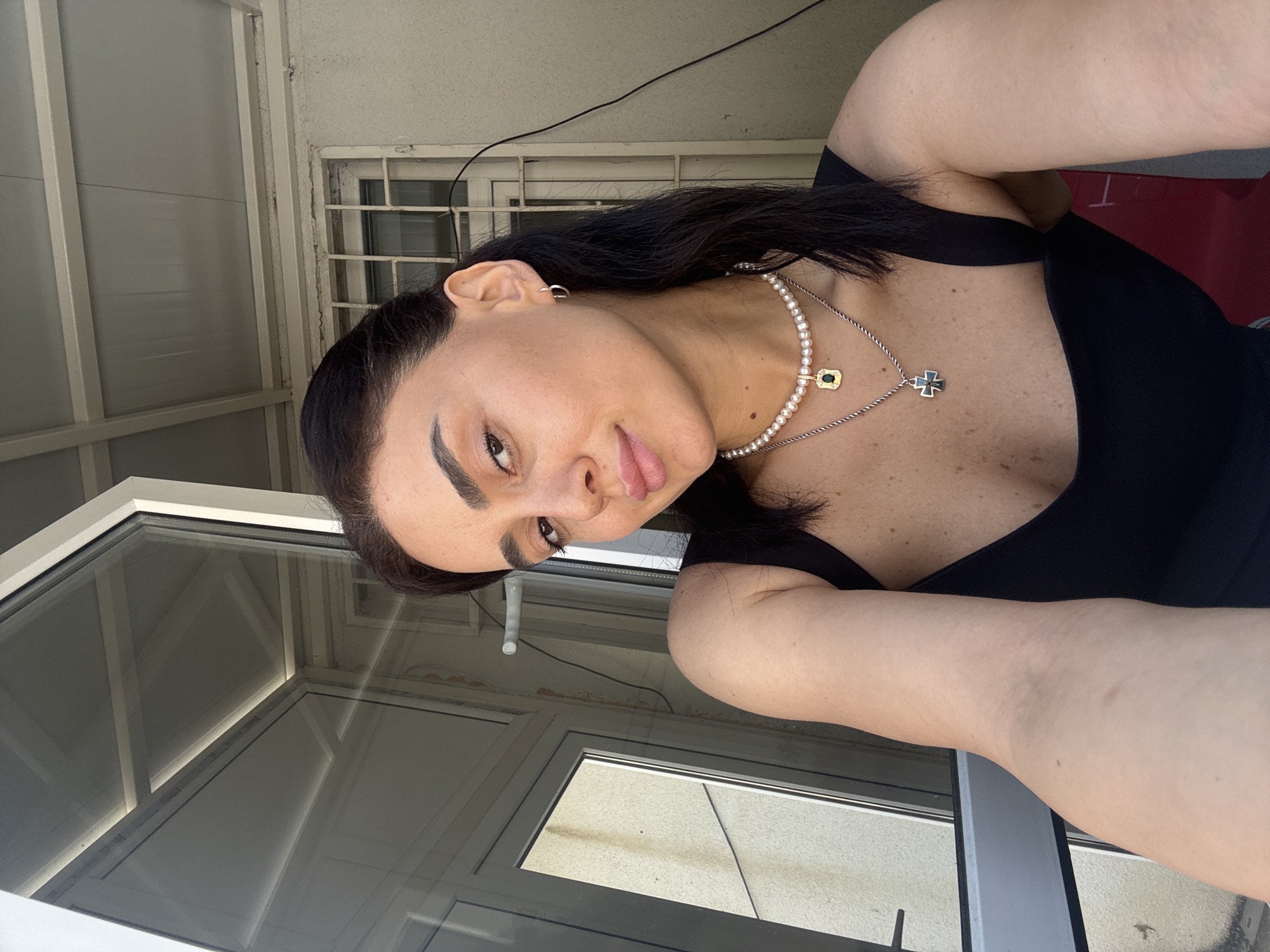 A woman with dark hair, wearing a black top and pearl necklaces, poses for a selfie by an open glass door. The background shows a building with barred windows and a corrugated metal roof, suggesting an urban setting possibly in Tbilisi, Georgia. The lighting indicates it's daytime.