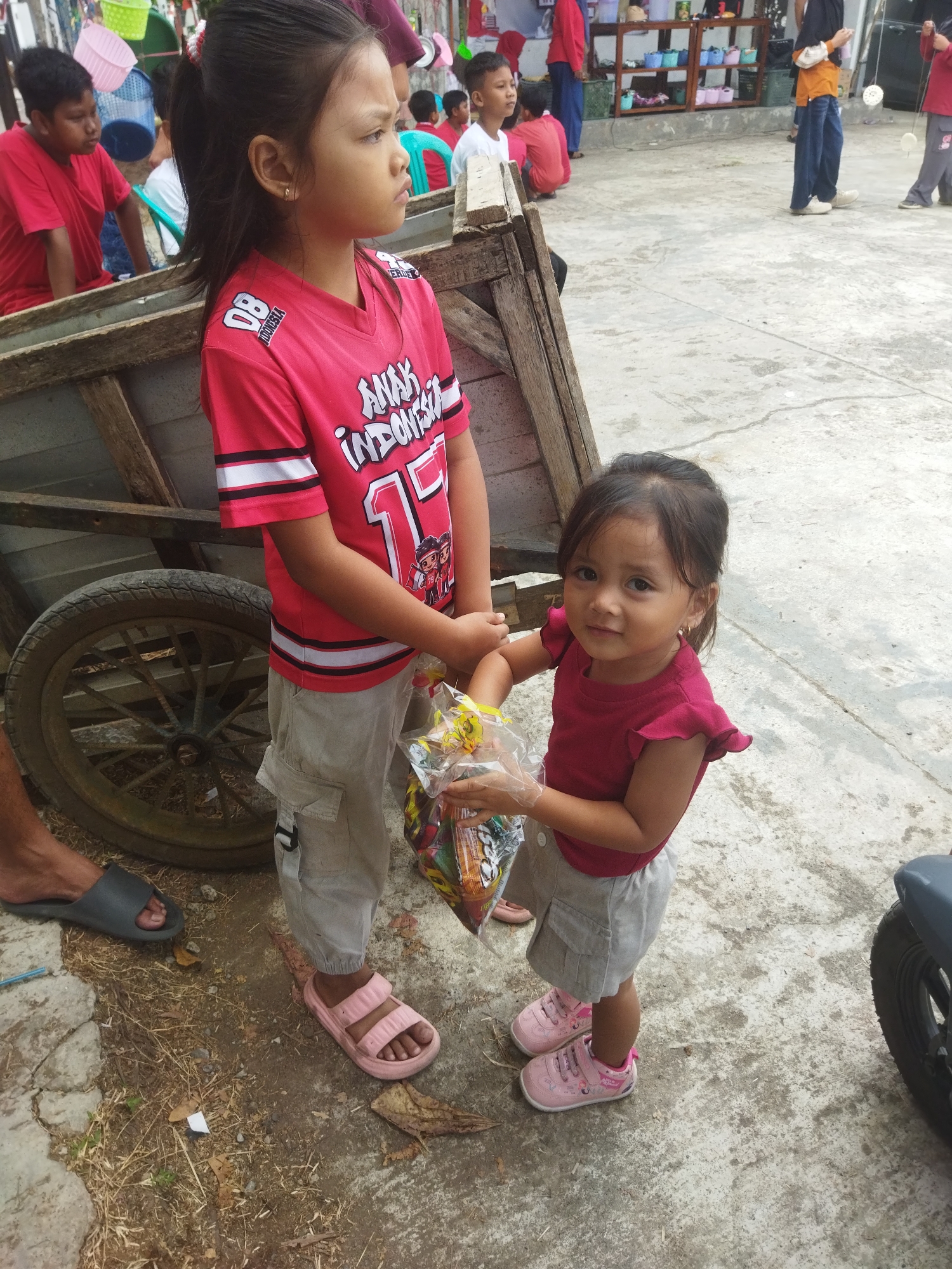 In Banjar, Indonesia, a young girl in a pink shirt and grey shorts stands near a wooden cart. She holds a bag of snacks. Next to her, an older girl in a red shirt and grey cargo pants also holds the snack bag. In the background, other people are visible, some sitting, and a market stall is set up with various colorful baskets. The ground is paved with concrete and dirt. A motorcycle wheel is partially visible on the right side of the frame.