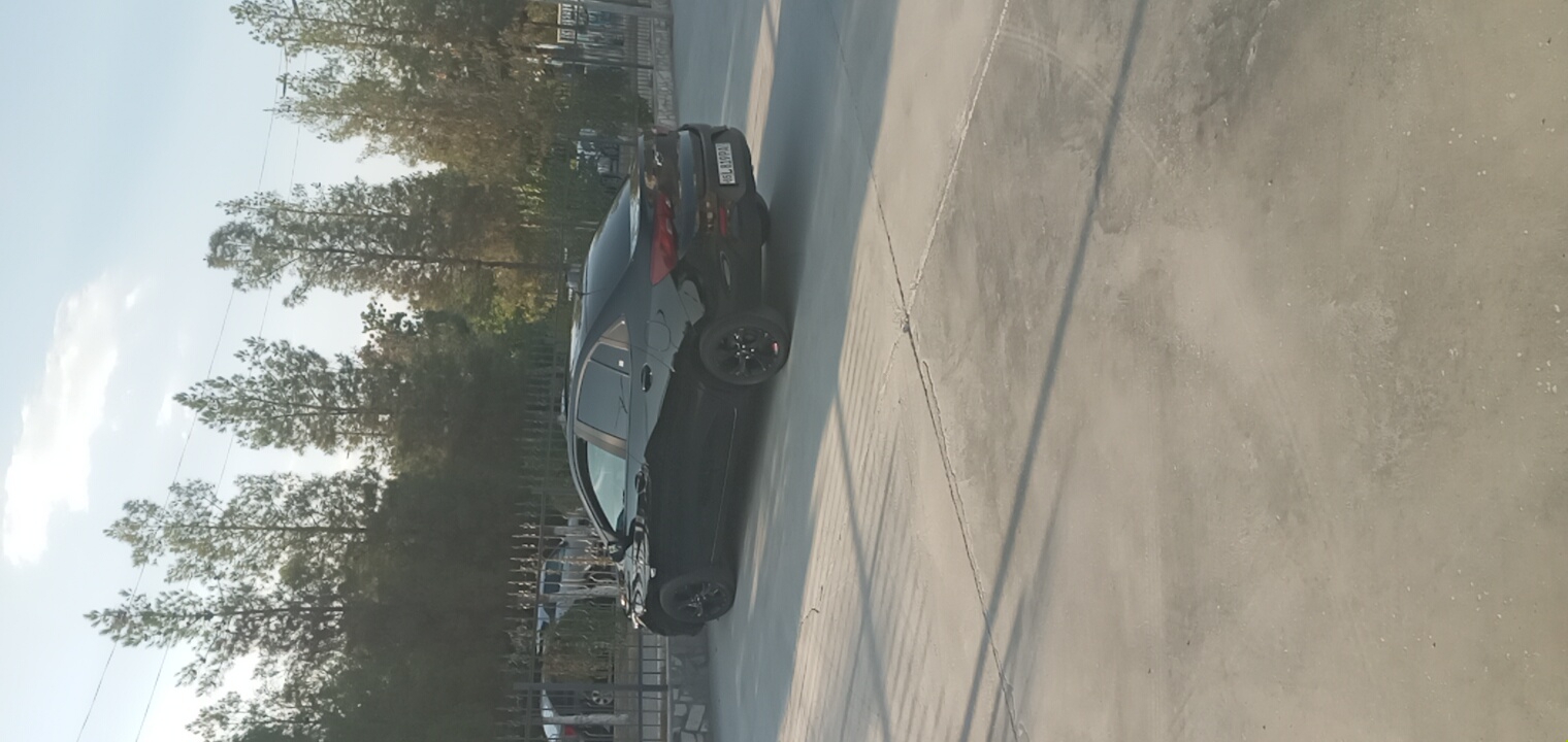 A black sedan is parked on a concrete lot with tire marks and shadows visible on the ground. In the background, there is a black metal fence, trees, and buildings. The sky is blue with some clouds. The license plate of the car reads "4SL819PA". The scene is consistent with an outdoor parking area in Navoiy City, Uzbekistan, possibly near a commercial or residential building given the fence and trees.