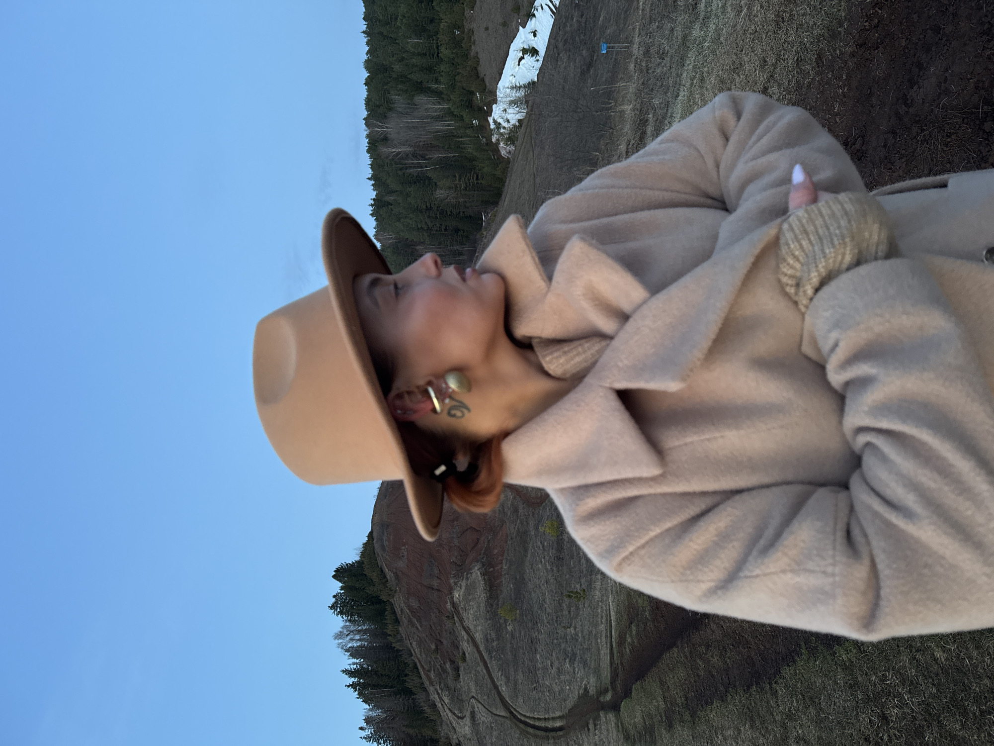 A woman wearing a beige coat and hat stands outdoors with her arms crossed in front of her. She is facing the sky and her head is tilted slightly upwards. She has earrings and a ring on her finger. Behind her, there is a hillside with trees and some snow. The image was taken in Ольховка, Russia.