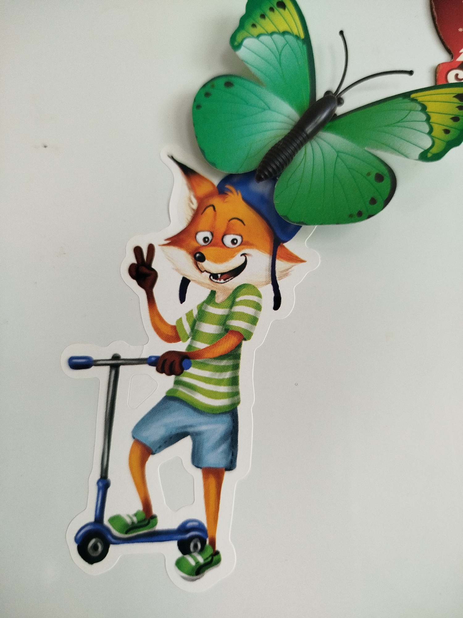 A sticker of a cartoon fox on a scooter is shown, with a green butterfly sticker behind it. The fox is wearing a blue cap, a green and white striped shirt, and blue shorts. It is holding up two fingers in a peace sign with its right hand and holding the handlebars of the scooter with its left hand. The scooter is blue and has black wheels. The background is a plain white surface. This image does not depict any specific location in Kryvaselski Sel'ski Savet, Belarus.