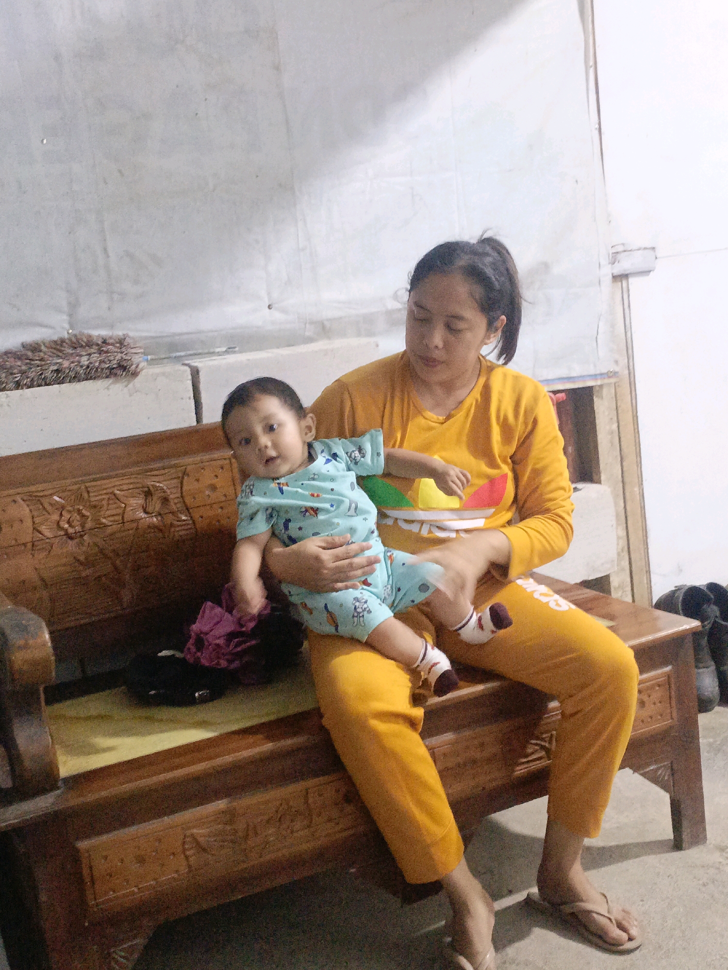 In an indoor scene, a woman is seated on a wooden bench, holding a baby. The woman, with her hair tied back, wears a yellow long-sleeved shirt with a colorful Adidas logo on the chest and yellow pants. The baby, dressed in a light blue outfit with cartoon characters, is held in her arms, facing forward. The wooden bench has intricate carvings. On the bench beside them are a dark object and a crumpled purple cloth. The background features a plain, light-colored wall with some texture and a faint smudge. To the right of the bench, a pair of dark boots are visible. The floor is a light-colored concrete. The overall impression is a domestic setting, likely a home interior. The specific location within Pager, Indonesia, cannot be determined from the visual information provided.