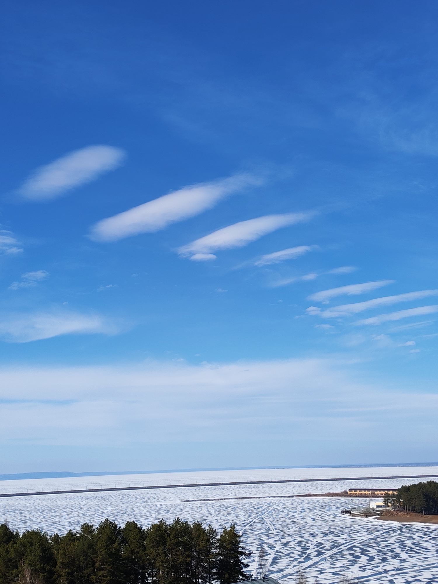 The image shows a winter landscape, likely in Chaikovsky, Russia. The sky is a bright blue with scattered cirrus clouds arranged in parallel lines, especially in the upper half. Below the sky is a large body of water covered in ice and snow. Tracks are visible on the ice surface, suggesting vehicular or pedestrian traffic. A dark, narrow strip runs across the frozen body of water, perhaps a road. Adjacent to the body of water is a landmass covered with a dense evergreen forest. On a small promontory jutting into the water is a small building complex, with light colored structures among the trees. A distant horizon line, indicating more land, is barely visible in the background. The entire scene evokes a sense of cold and open space.