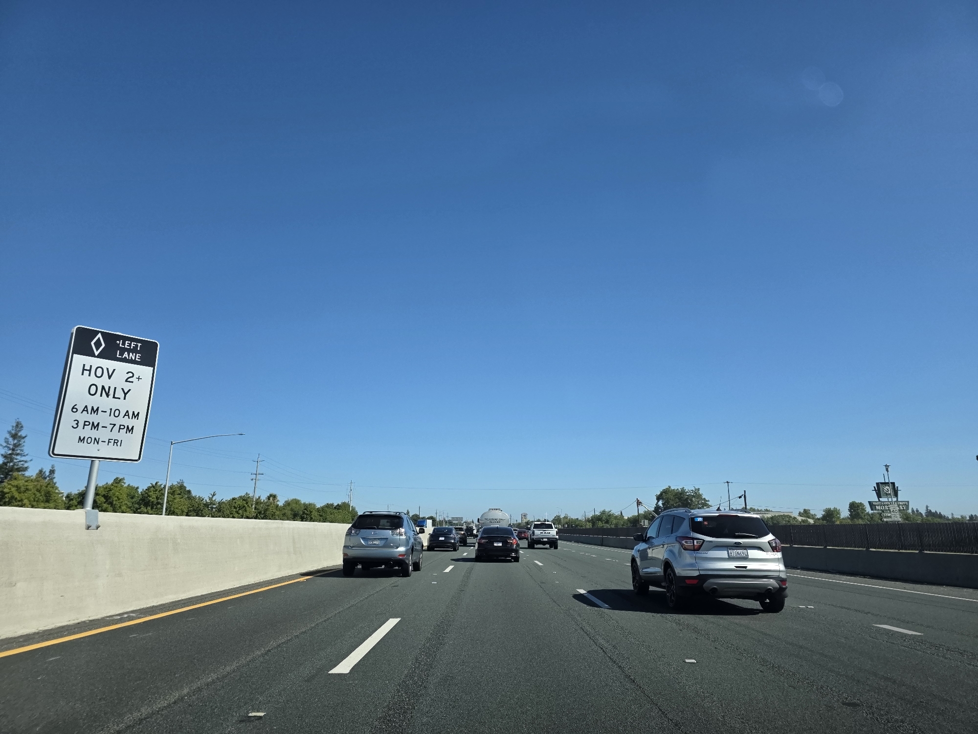 A view from inside a vehicle traveling on a highway. The sky is clear and blue. A concrete barrier runs along the left side of the road, and a diamond-shaped sign indicates "LEFT LANE HOV 2+ ONLY" with specific hours of operation. Several cars are traveling in the same direction on the highway, including a silver SUV in the foreground, a dark sedan, and a white truck. Trees and power lines are visible in the background, along with a distant sign structure. The road is asphalt with white lane markings. The license plate of the silver SUV is visible and reads "8TGW694". The scene appears to be in Elk Grove, California, United States of America, based on the context of the HOV lane sign common in that region.