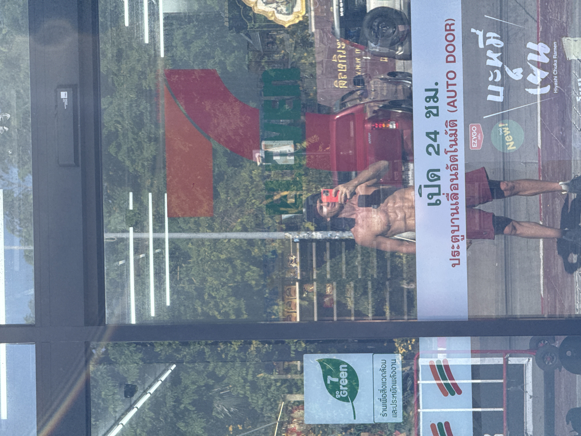 A shirtless man with dark hair and a bandana is taking a selfie in the glass reflection of a 7-Eleven store. He is wearing red shorts and black athletic shoes. The store's interior is visible through the glass, showing shelves stocked with products and fluorescent lighting. Outside, trees and a red pickup truck are reflected. A sign on the door indicates "7 Green" with Thai text. Another sign above the door reads "OPEN 24 HRS. AUTOMATIC DOOR." Text in Thai script is also visible on the door, possibly indicating food items, with "Hiyashi Chuka Ramen" printed in English at the bottom. The man is positioned in the foreground, with the store and its surroundings reflected around him. The scene appears to be in ตำบลเกาะพะงัน, Thailand.