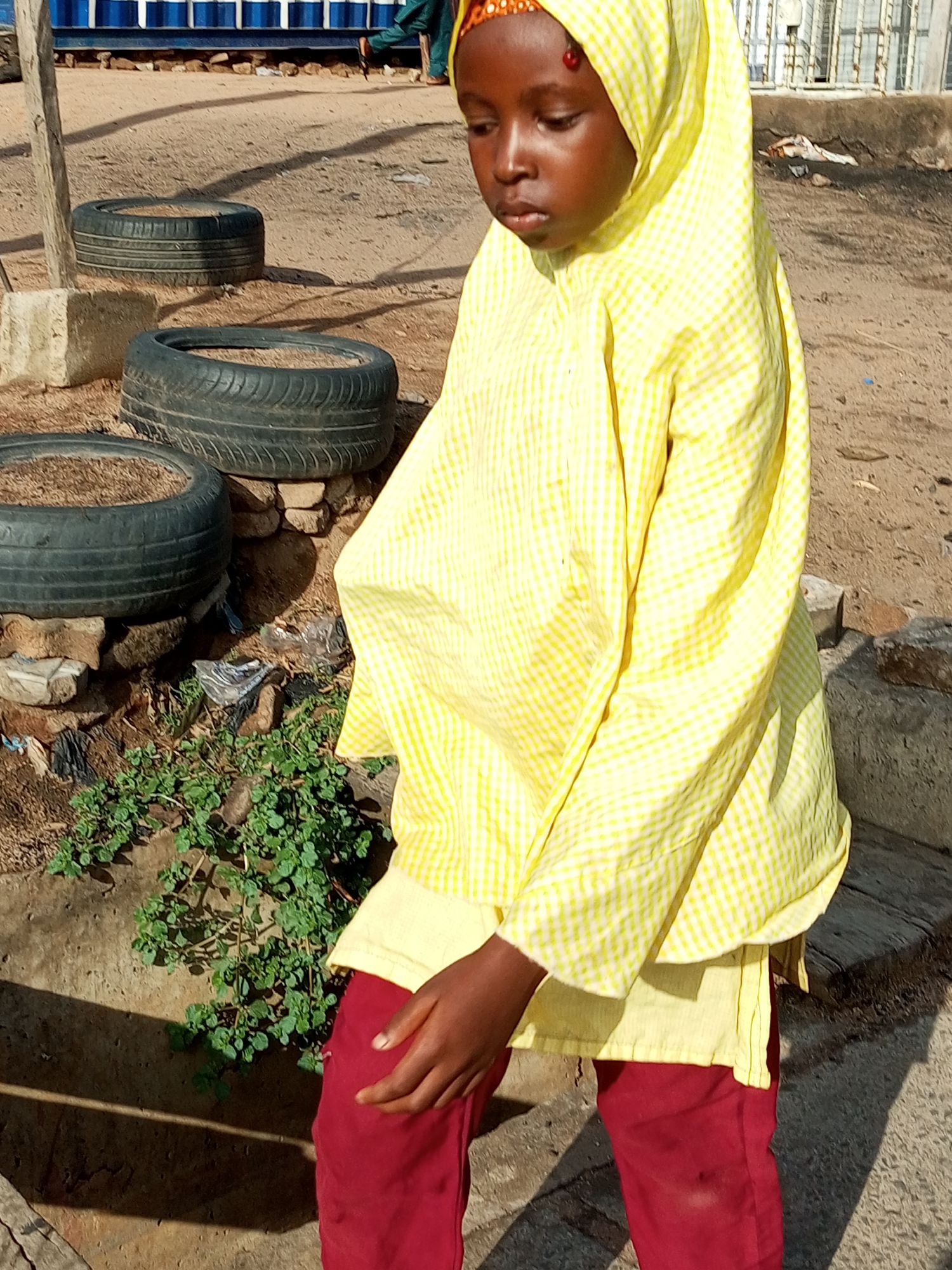 A young girl, possibly of Nigerian ethnicity, stands outdoors in Garko, Nigeria. She wears a yellow checkered hijab and a matching long-sleeved shirt, paired with dark red pants. Her right hand rests on her hip. The background consists of a dusty, sandy ground, with several discarded tires arranged in rows. Some of the tires appear to be filled with dirt. Green foliage grows in front of a concrete barrier. In the distance, a blue and white structure with a corrugated metal roof is visible. The lighting suggests it is daytime, casting distinct shadows on the ground.