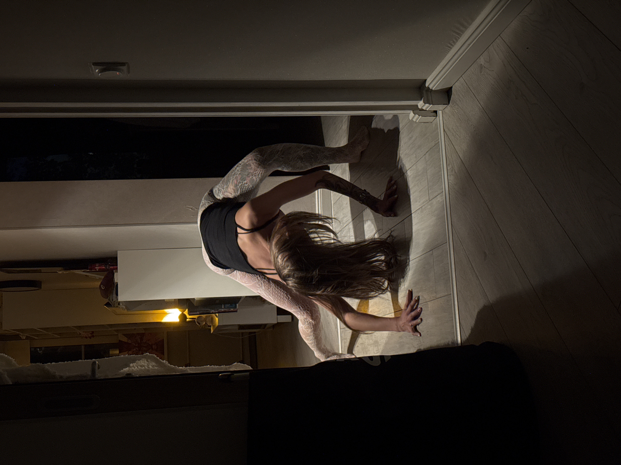 A person is performing a yoga pose in a hallway. The person is in a bridge pose, with their hands on the floor and their body arched. They are wearing a black tank top and white patterned leggings. The hallway has a wooden floor and is dimly lit. There are doors and furniture visible in the background. The image does not contain any information that can be used to determine the location in Moscow, Russia.