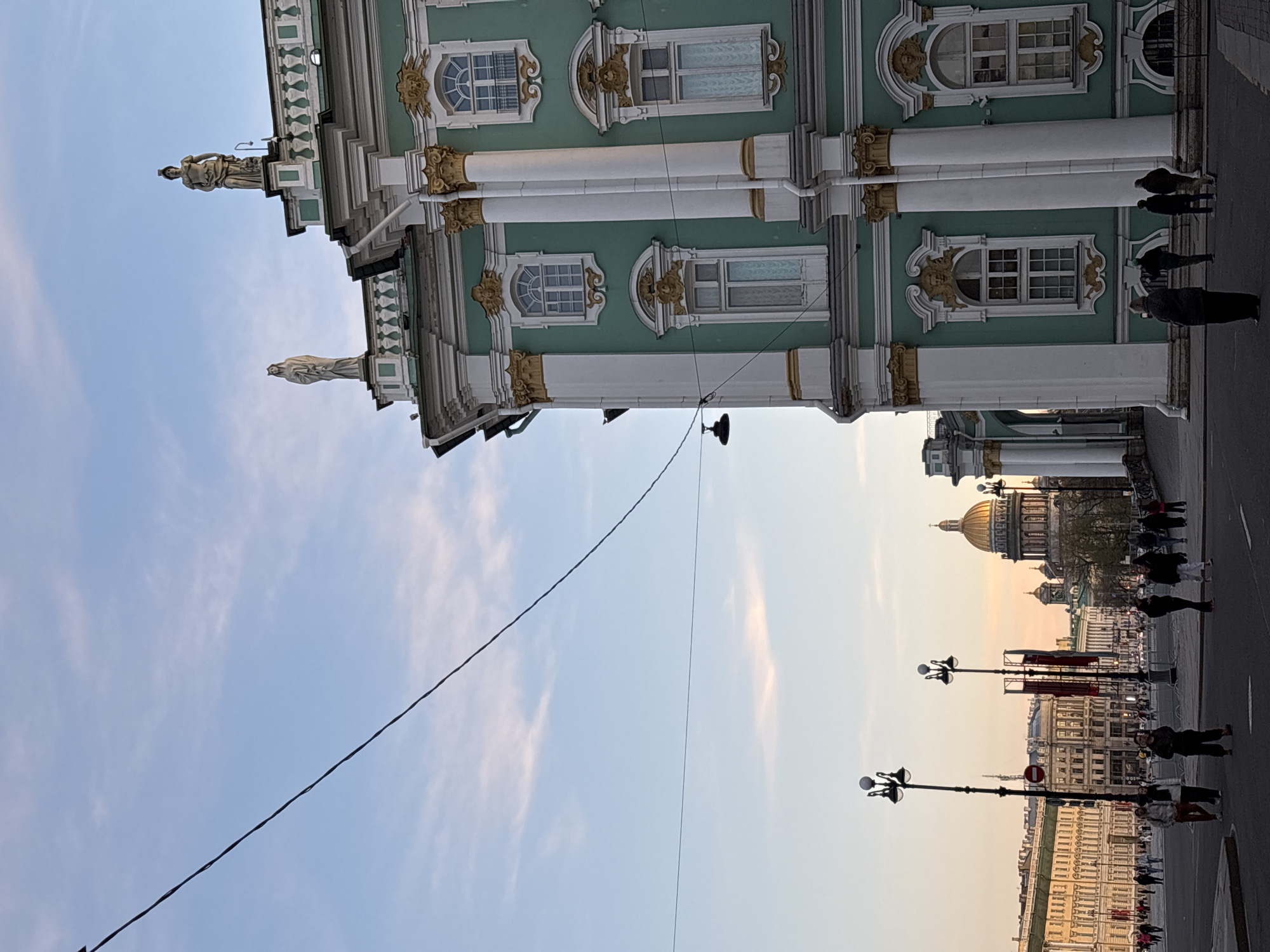 The image captures a street view in Saint Petersburg, Russia, featuring the Winter Palace (part of the Hermitage Museum complex) on the left. The palace is identifiable by its distinctive mint-green facade, ornate white columns, golden detailing, and classical statues. On the right side of the frame, across a street filled with pedestrians, is the distinctive golden dome of St. Isaac's Cathedral. The sky above is a gradient of blue, fading to a light orange near the horizon. Overhead power lines intersect the view. Street lamps are visible lining the street. Pedestrians are walking on the street.