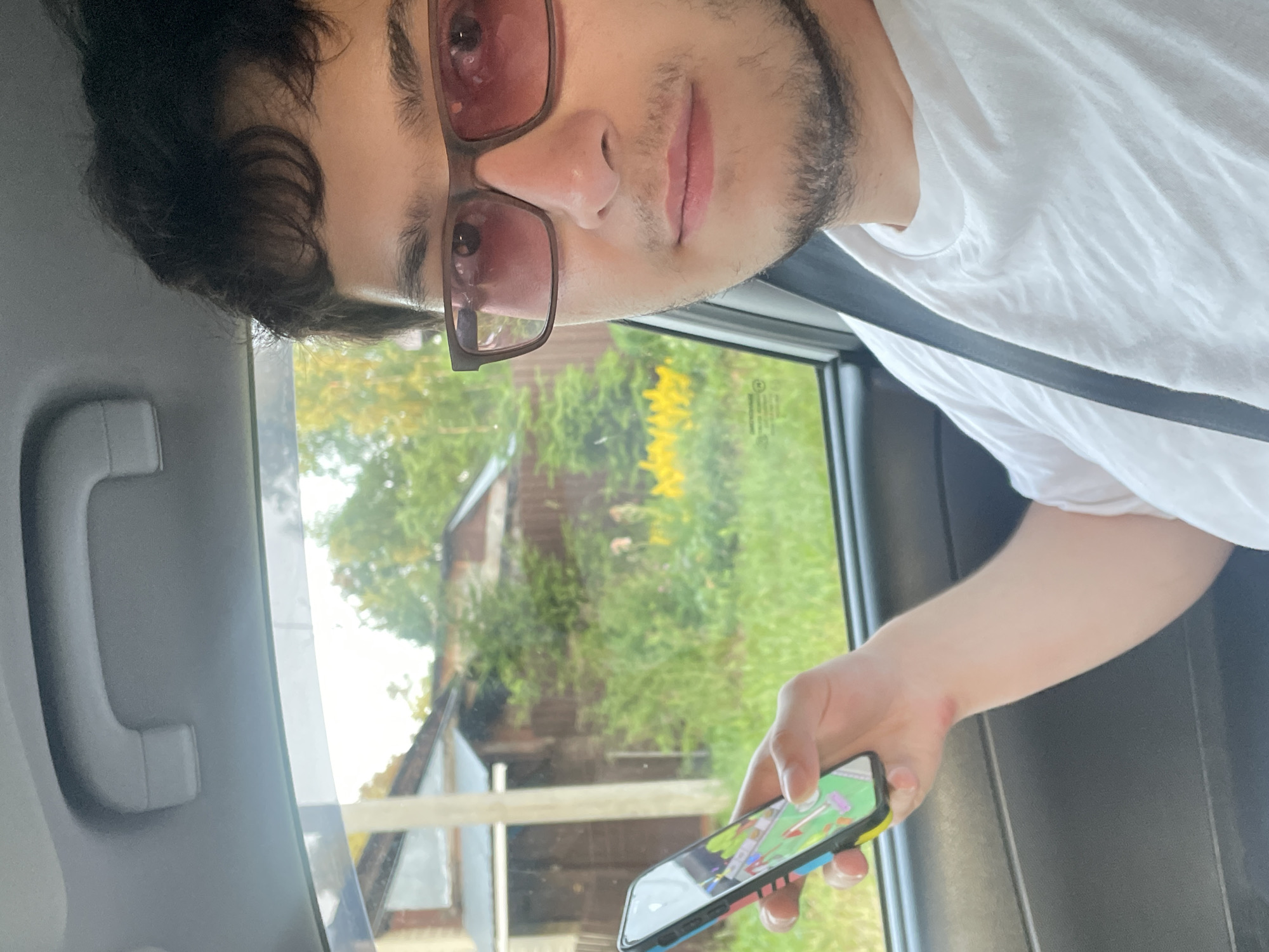 A person, wearing a white t-shirt and pink-tinted glasses, is sitting in the passenger seat of a car and holding a smartphone. The smartphone displays a game with colorful graphics. The car's interior is visible, including a grab handle on the ceiling and a seatbelt. Outside the car window, a suburban or rural landscape is visible, with houses, trees, and yellow flowers. The scene appears to be set in the city of Votkinsk, Russia. The lighting suggests it is daytime. The person is looking towards the camera.