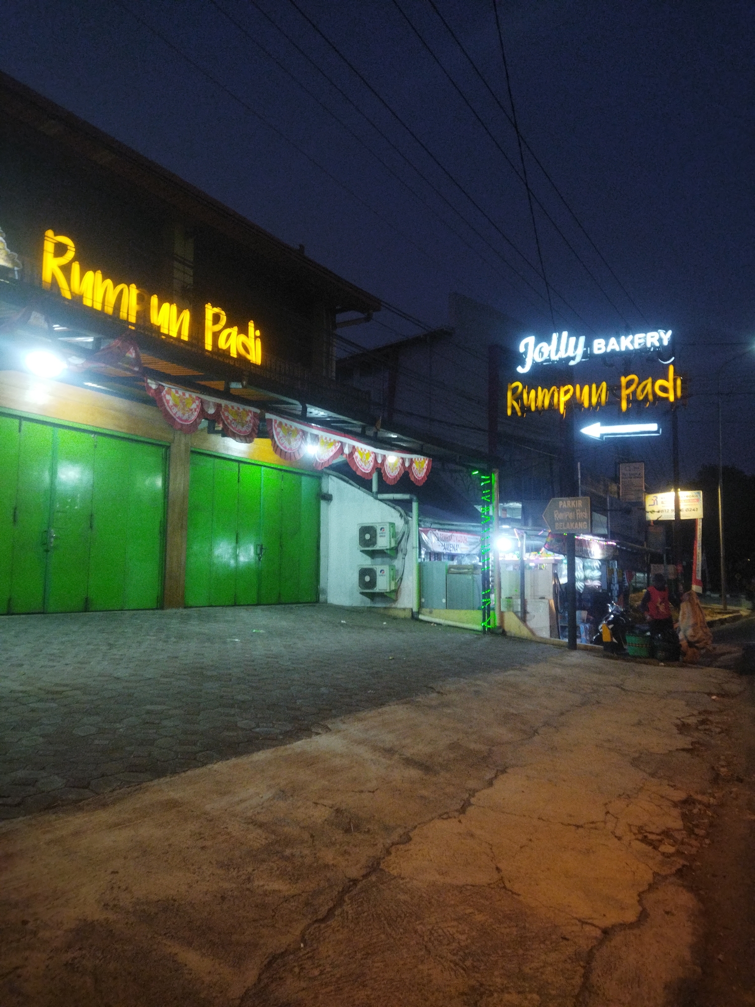 An exterior, eye-level shot taken at night shows a row of shops in Wonogiri, Indonesia. The sky is dark blue with visible power lines crisscrossing the frame. A yellow neon sign with the text "RUMPUT UN PADI" is prominently displayed on the left side of the image. To the right of this, a blue neon sign reads "JOLLY BAKERY," with "RUMPUN PADI" below it in yellow neon. A white neon arrow points to the left from the "RUMPUN PADI" sign. In front of the shops are two large, vibrant green metal roller doors. Above these doors, a decorative banner with red and white patterned flags is strung across. Two white air conditioning units are mounted on the wall to the right of the green doors. The ground in front of the shops is paved with interlocking stones, and further in front, the ground is cracked concrete. Several people are visible in the background, including two individuals in the far right, one wearing a red top and another in a beige garment. A motorcycle is parked on the right side of the frame. A sign with text in Indonesian, including "PARKING," is also visible.