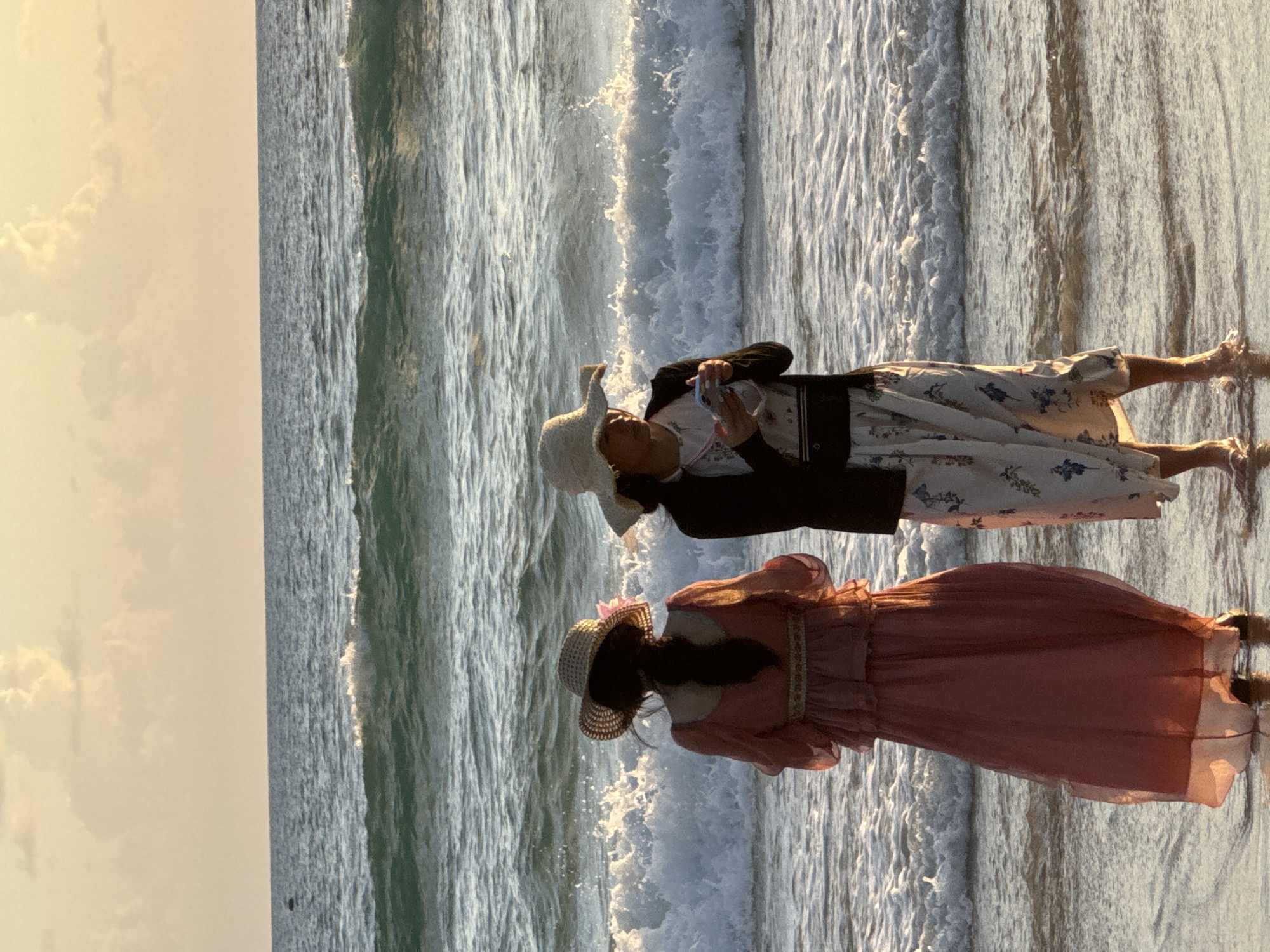 Two individuals, viewed from the rear, stand on a wet sandy beach facing the ocean. The person on the left wears a long, sheer pink dress with tiered ruffles and a wide-brimmed straw hat adorned with a pink flower. The person on the right wears a floral print dress with a white base and blue flowers, a black cardigan, and a wide-brimmed straw hat. They are holding a phone, seemingly taking a picture or recording. Gentle waves are breaking on the shore, with a spray of foam visible in the mid-ground. The sky above is a soft gradient of pale yellow and orange, indicative of either sunrise or sunset, with a few scattered clouds. The location is Karon Beach, Thailand, characterized by its calm, serene waters and picturesque sunset sky.