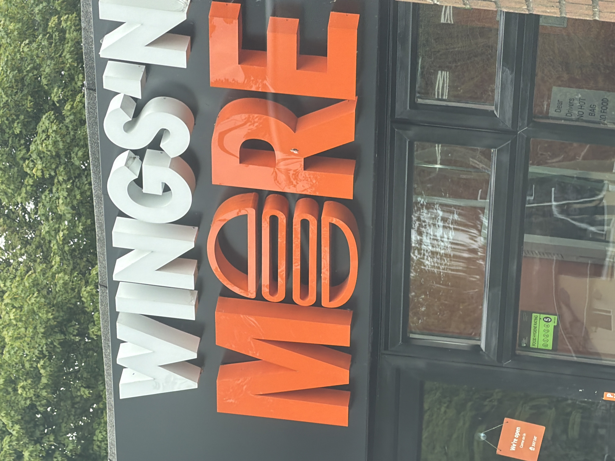 The image depicts a storefront in Pity Me, United Kingdom. A sign reading "WINGSIN MORE" is prominently displayed, with "WINGSIN" in white letters and "MORE" in orange. A window next to the sign has stickers and signs, including a food hygiene rating of 5, a note to delivery drivers, and an "open" sign advertising Just Eat. Foliage of a tree is visible in the background. The location appears to be outdoors.