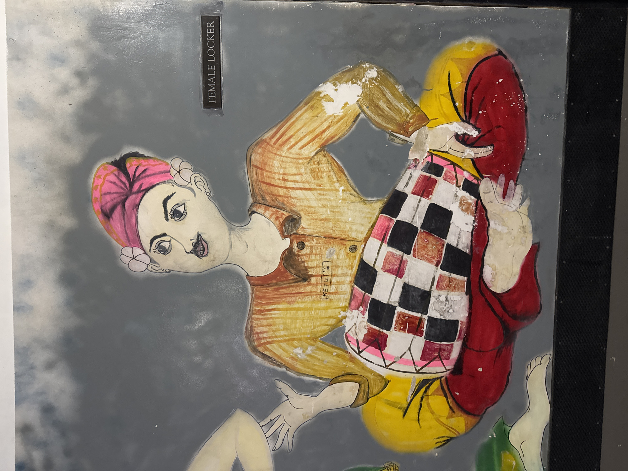 An oil painting on a grey background depicts a figure with a pink turban, a yellow and orange striped shirt, and red pants, playing a drum. The figure is sitting cross-legged, with its hands on the drum. The drum has a checkered pattern of black, white, and red squares. To the right of the figure, a dark rectangular sign reads "FEMALE LOCKER". The painting appears to be on a wall in a public space, possibly a locker room, given the sign. The artwork shows signs of wear and tear, with some paint peeling off. The overall style is illustrative and somewhat naive. The painting is located on a wall in Kuta Selatan, Indonesia.