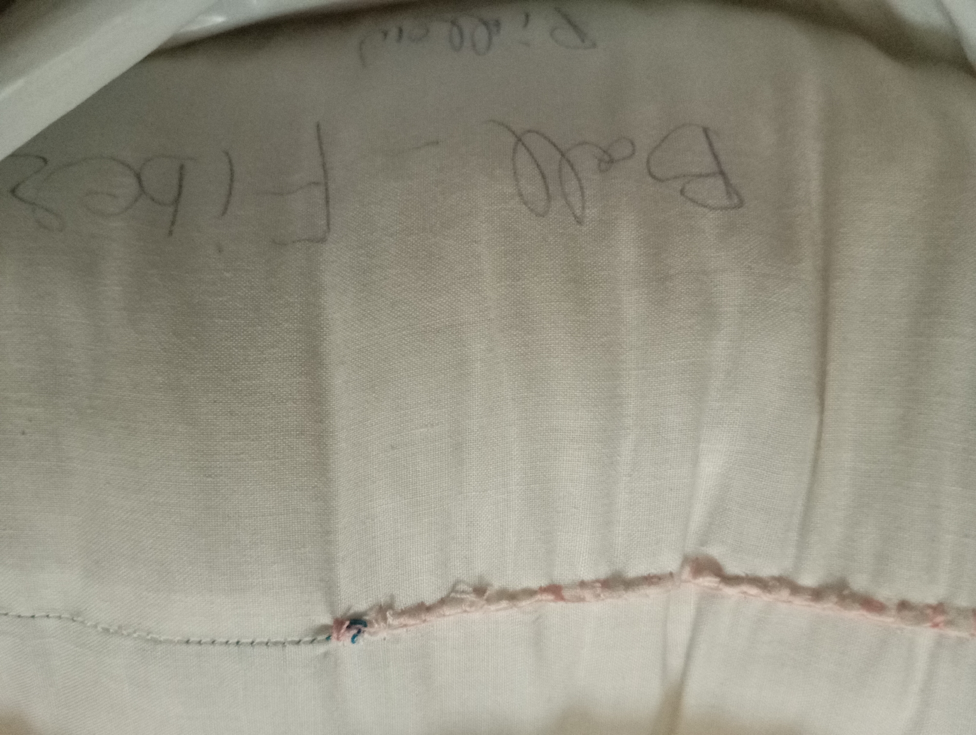 The image shows a close-up of a light-colored fabric, possibly a pillowcase or upholstery. Hand-written text in dark pencil is visible on the fabric. The text appears to read "Fiber Ball Pillow". There are also some small, colorful threads or stitches visible along a seam or edge of the fabric, possibly indicating repair or decoration. The lighting suggests the photograph was taken indoors. No specific location in a city or any people are depicted in the image.