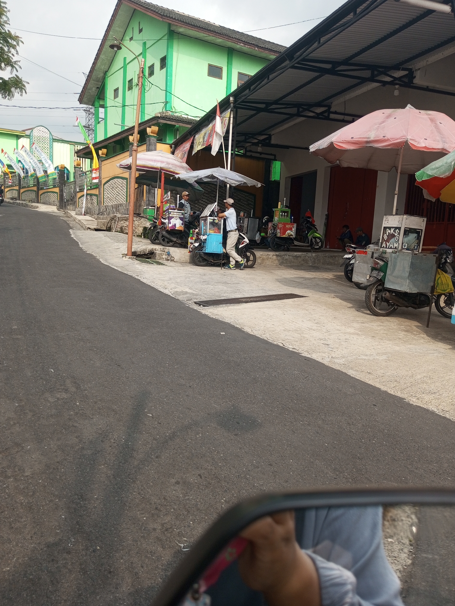 The image depicts a street scene in Krasak, Indonesia. The foreground is dominated by a road with a visible shadow, and a motorcycle's side mirror reflects a person's hand and arm. In the midground, a row of food carts with umbrellas are set up along a sidewalk. One cart displays text that appears to be "BAKSO AYAM" which translates to "chicken meatballs". Several people are attending to the carts or are in the vicinity. A green building with a tiled roof is visible behind the food carts, and flags are displayed along the street. The environment suggests a commercial area with street vendors.
