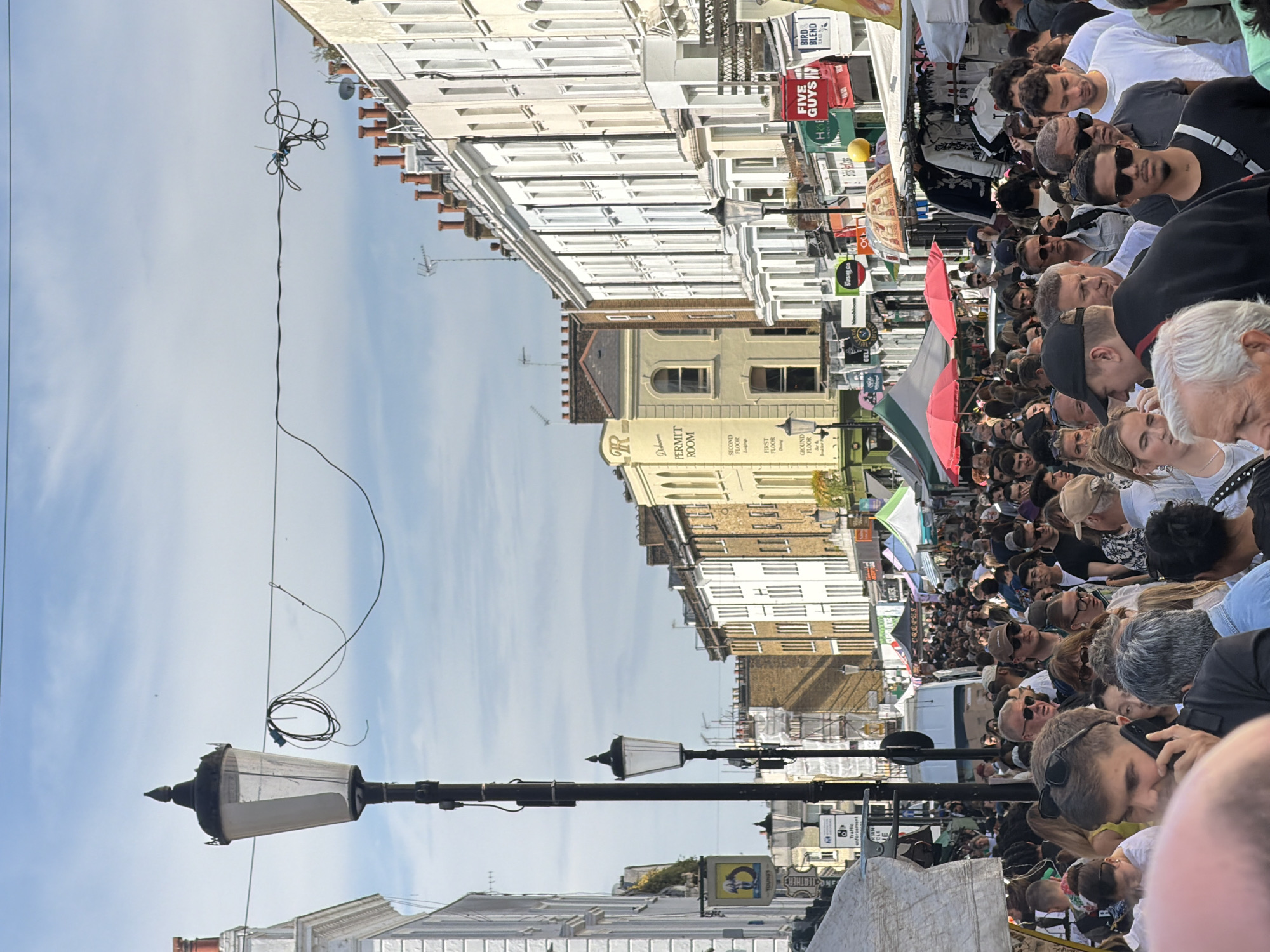 The image captures a crowded street scene in London, United Kingdom. The focus is on the dense crowd of people filling the street, suggesting an event or market is taking place. Buildings line both sides of the street, showcasing various architectural styles, including white-fronted buildings, brown-brick buildings, and structures with distinctive signage like "PERMIT ROOM." Street elements include a lamp post and overhead wires. Flags and market stalls are visible within the crowd. Signs for "FIVE GUYS," "BIRD BLEND TEA CO," "SUSHI.CO," "Traffic," "LEATHER," and "CREPES" are discernible among the commercial establishments.