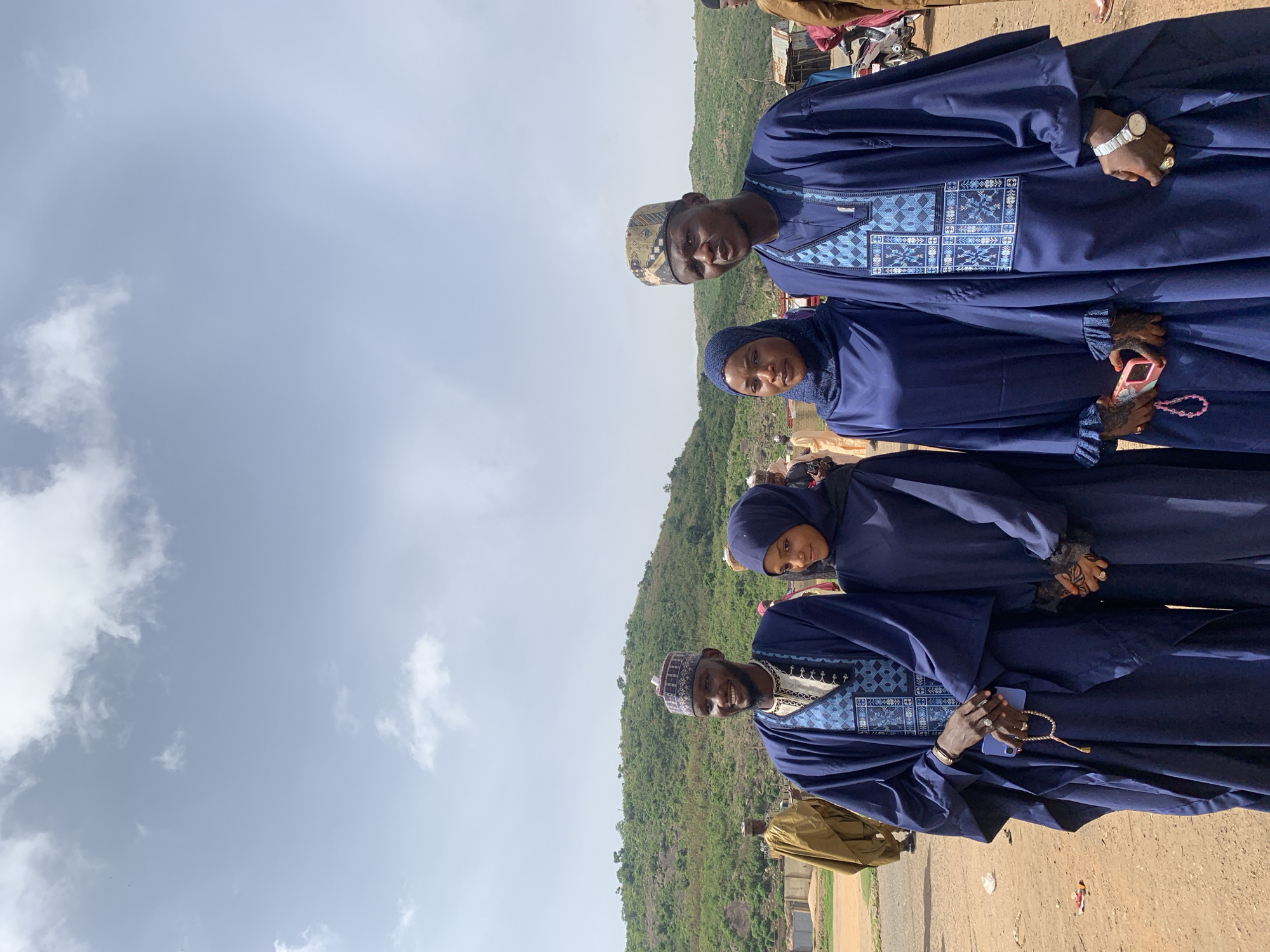 Four individuals are captured in an outdoor photograph in Naraguta Hausawa, Nigeria, taken during daylight under a partly cloudy sky. Two men, positioned at the top and bottom of the frame, and two women, located in the middle, are all wearing similar, flowing blue garments. The men have matching hats, while the women have head coverings. The men are holding mobile phones and prayer beads, and one woman holds a pink smartphone. The background features a green, hilly landscape and a dirt road. Other people and vehicles are blurred in the background.