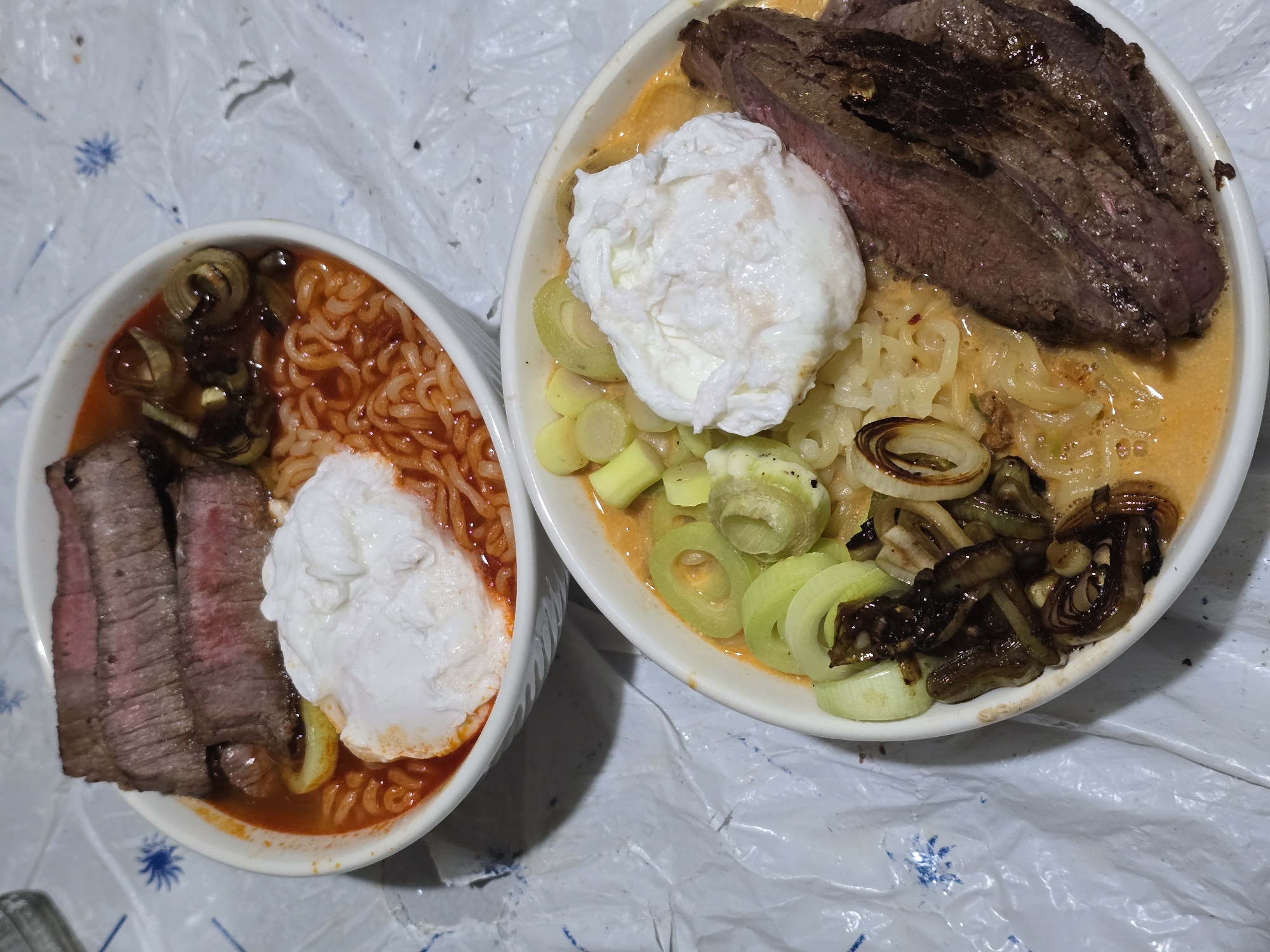 The image shows two bowls of food, likely ramen or noodle soup, on a white plastic surface. Each bowl contains noodles, broth, sliced meat (possibly steak), a poached egg, and sliced vegetables (possibly scallions or leeks). The left bowl has red-orange broth and the right bowl has yellowish broth. There are no people visible in the image. The location is not identifiable from the image. The environment is not identifiable from the image. The city of Sofia, Bulgaria is not identifiable from the image.