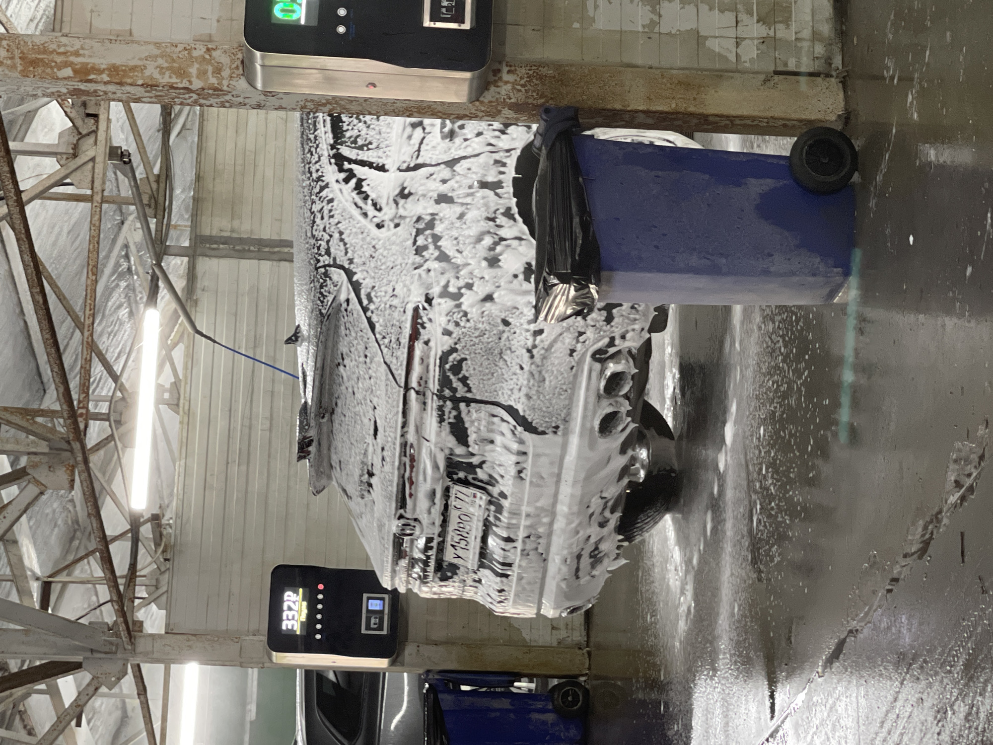 An indoor car wash bay is depicted, where a dark-colored car is actively undergoing a wash cycle. The vehicle, seen from its rear-left, is entirely covered in a thick layer of white cleaning foam, indicating it is in the foaming stage of the wash. Its rear license plate, "У 150 РО 77", is visible through the suds, with the "77" region code confirming the location as Moscow, Russia.
To the right of the car, a blue, wheeled industrial trash can with a black plastic bag attached is positioned. Above and around the car, the utilitarian structure of the car wash is visible, featuring exposed, possibly rusty, metal beams, white paneling or insulation on the walls, and bright fluorescent lights illuminating the scene. A blue hose or wand extends from above towards the car.
Two digital display units are present. One in the upper part of the frame shows some green graphical elements and lights. Another unit in the lower left displays "33Zp" (or "332P") along with a sequence of indicator lights. The concrete floor is wet and reflects the bright artificial lighting, typical of a busy car wash environment. No individuals are visible in the image.